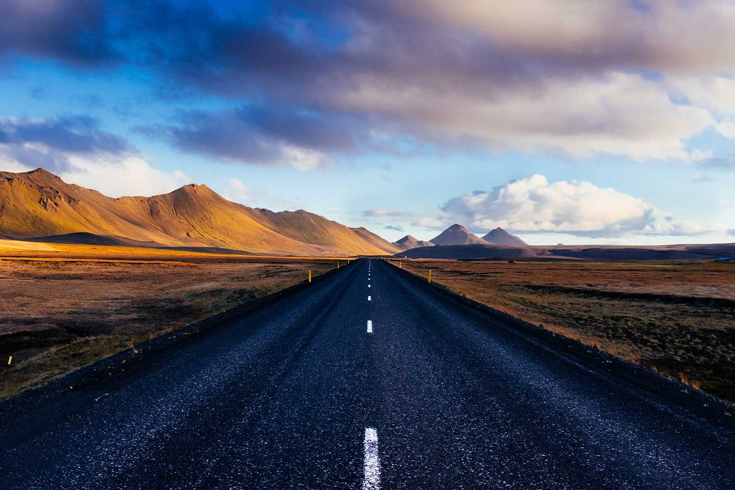 A long, straight road leads into a dramatic Icelandic landscape, bordered by golden-lit mountains on either side. The sky is filled with a mix of dark clouds and patches of blue, adding depth to the scene.