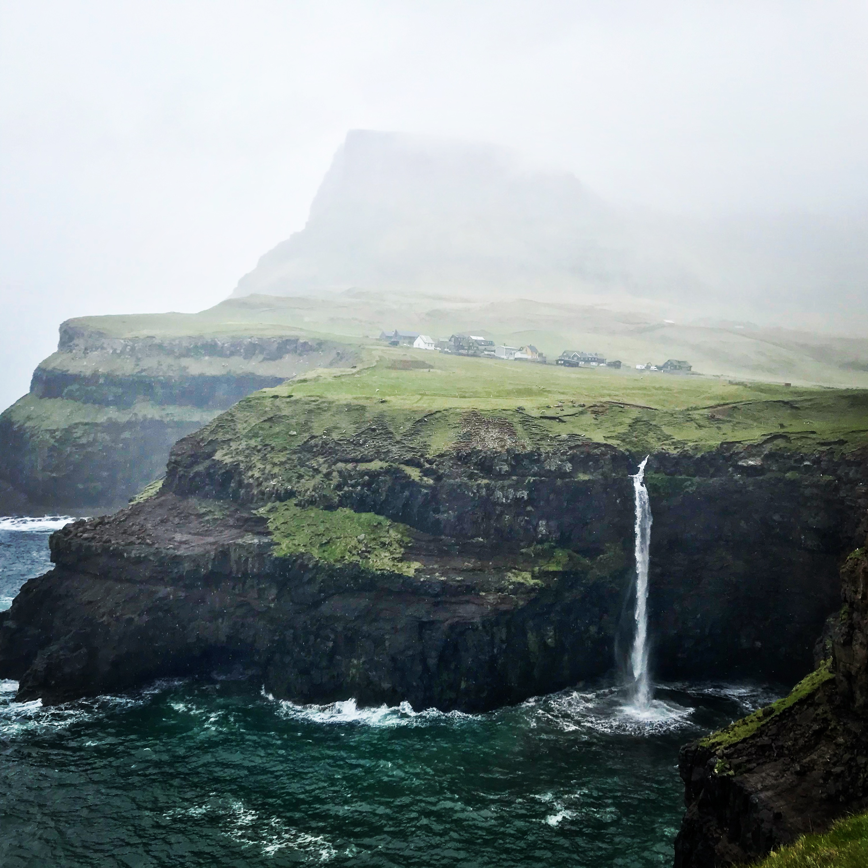 Gasadalur, Faroe IslandsThis image features a dramatic coastal scene with a waterfall plunging off a cliff into the ocean below. The misty atmosphere softens the distant view of a grassy plateau, dotted with a small cluster of houses, and a towering hill shrouded in fog. The dark cliffs and turquoise waters contrast beautifully with the lush green landscape, creating a serene yet rugged ambiance.