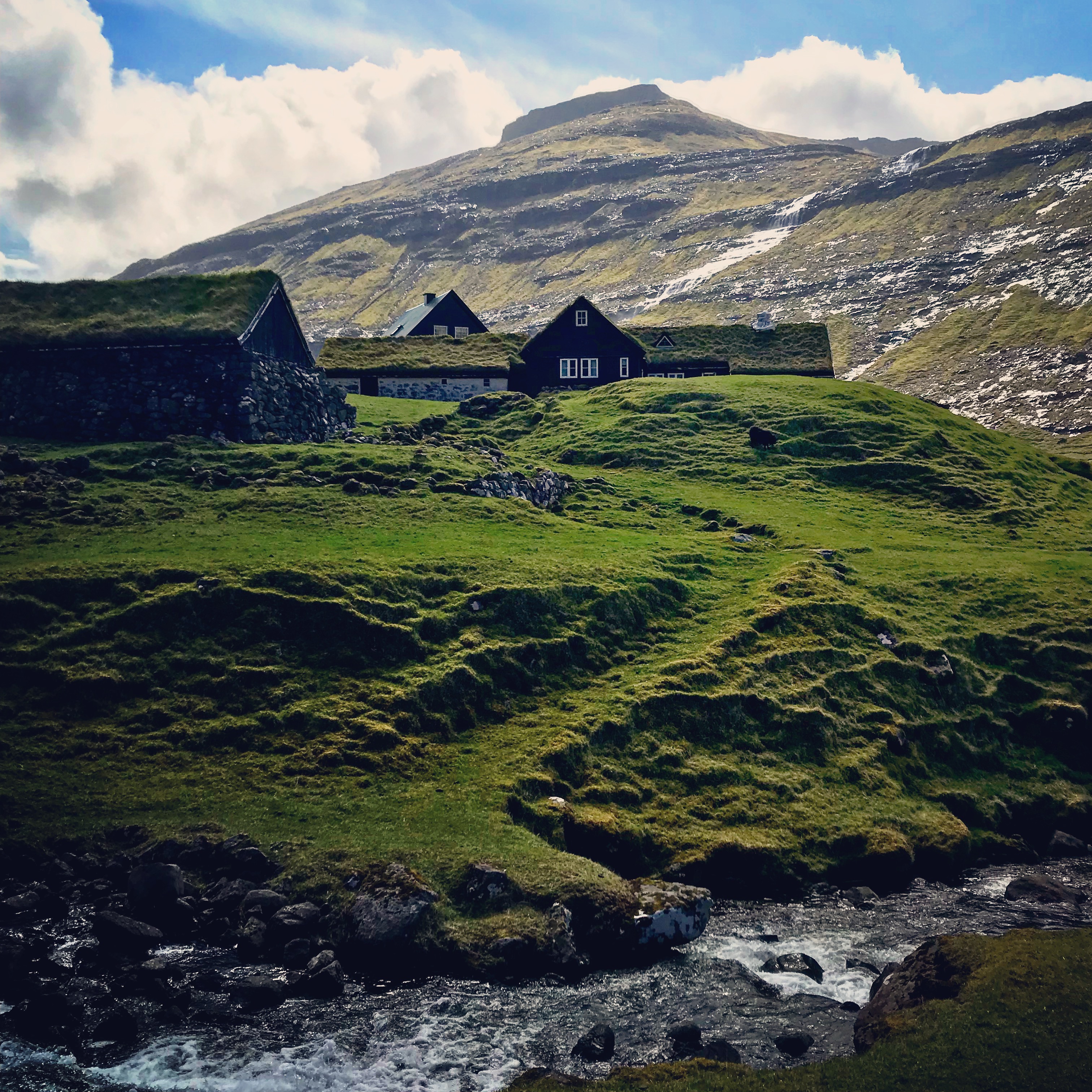 This image showcases a cluster of traditional turf-roofed houses set on a rolling green hillside in Saksun, with a clear stream winding through the foreground. The mountainous backdrop, partially covered with snow and illuminated by sunlight under a blue sky, adds a sense of scale and rugged beauty. The scene exudes a peaceful, pastoral charm, emphasizing the harmony between nature and architecture.