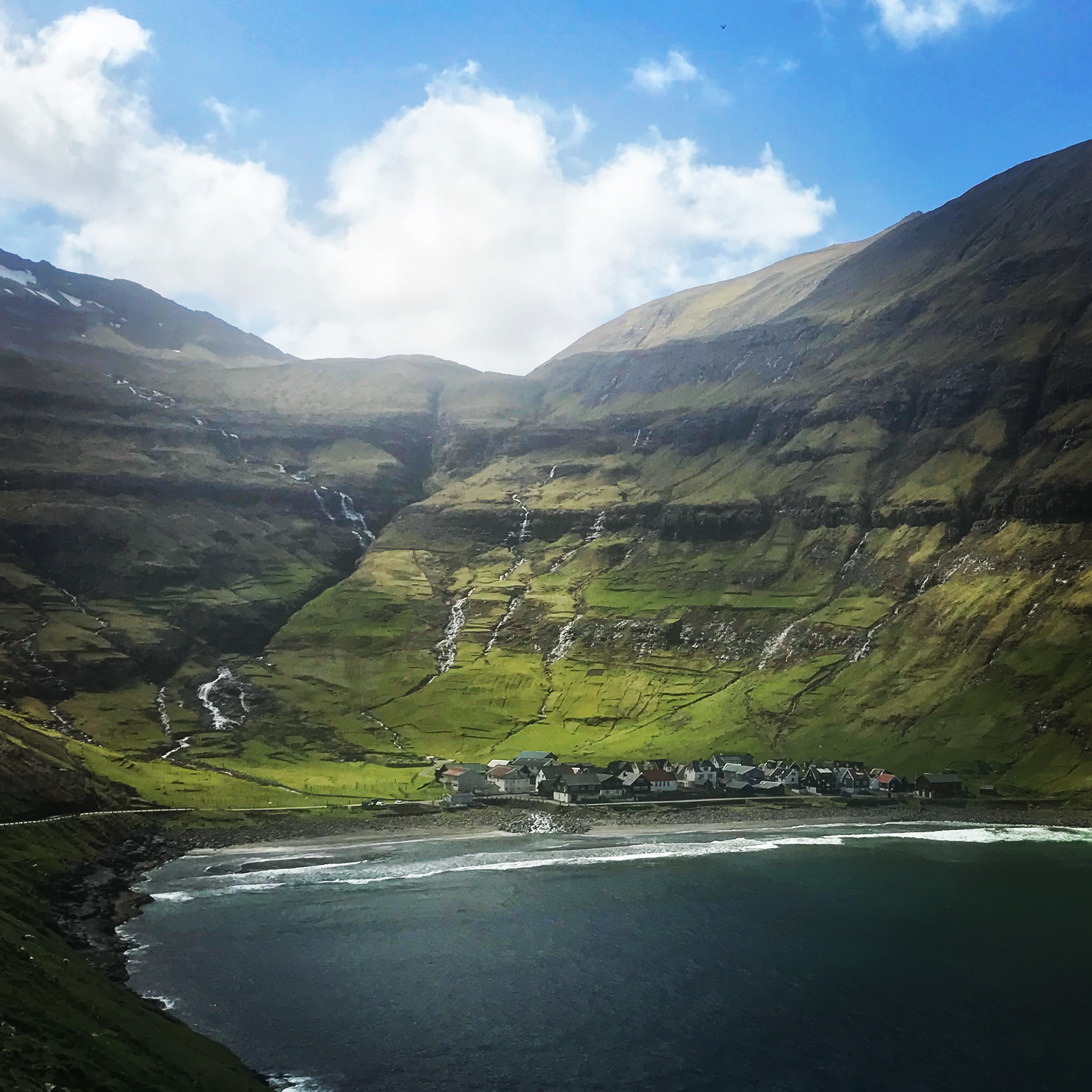 Tjornuvik, Faroe IslandsThis image depicts a small village nestled at the edge of a calm bay, surrounded by steep, green mountains with cascading waterfalls. The vibrant landscape is illuminated under a bright blue sky with scattered clouds, creating a serene and picturesque setting. The contrast between the water, rugged terrain, and quaint houses highlights the remote beauty of this location.