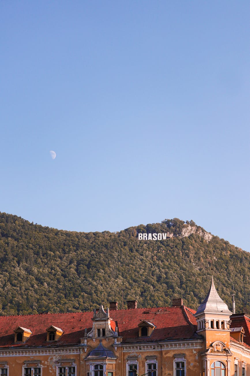This is an image of a picturesque view of a mountain with a forested slope featuring a "BRASOV" sign, reminiscent of Hollywood-style lettering, under a clear blue sky with a visible crescent moon. In the foreground, an ornate building with red roof tiles and detailed architecture complements the natural scenery.