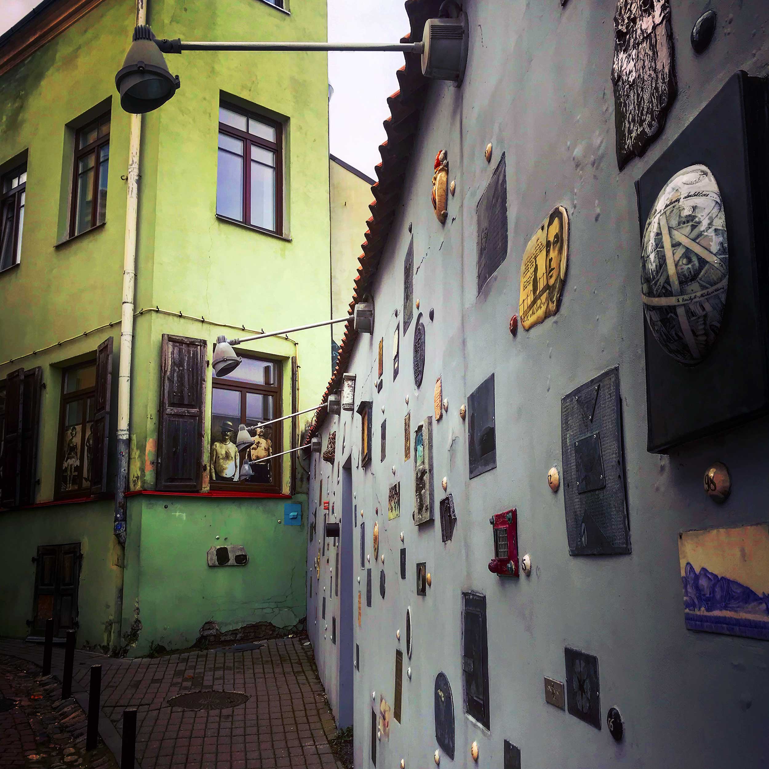 This image shows a narrow, artistic alley with a green building on one side and a unique wall installation on the other. The wall features an eclectic collection of mounted objects, including ceramic pieces, framed art, and relief sculptures, creating a whimsical outdoor gallery. The rustic windows of the green building add charm to the setting, making it feel like a hidden artistic corner.
