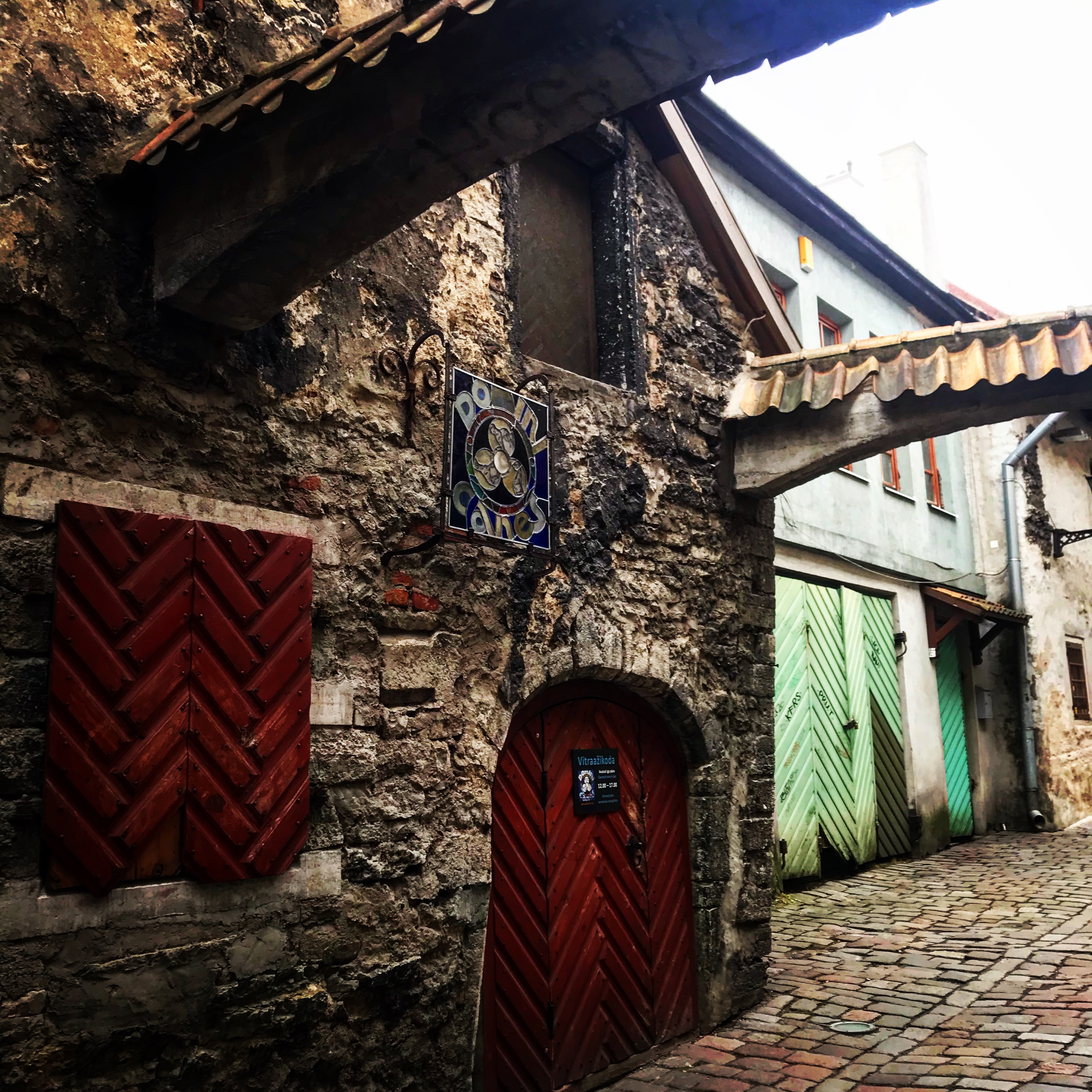 This image captures a narrow, medieval-style alley with weathered stone walls and red and green chevron-patterned wooden doors. An arched entrance with a small sign and a decorative emblem featuring the words "Poomi Kanes" adds a rustic charm. Overhead, tiled awnings connect the buildings, contributing to the cozy and historic ambiance of the scene.