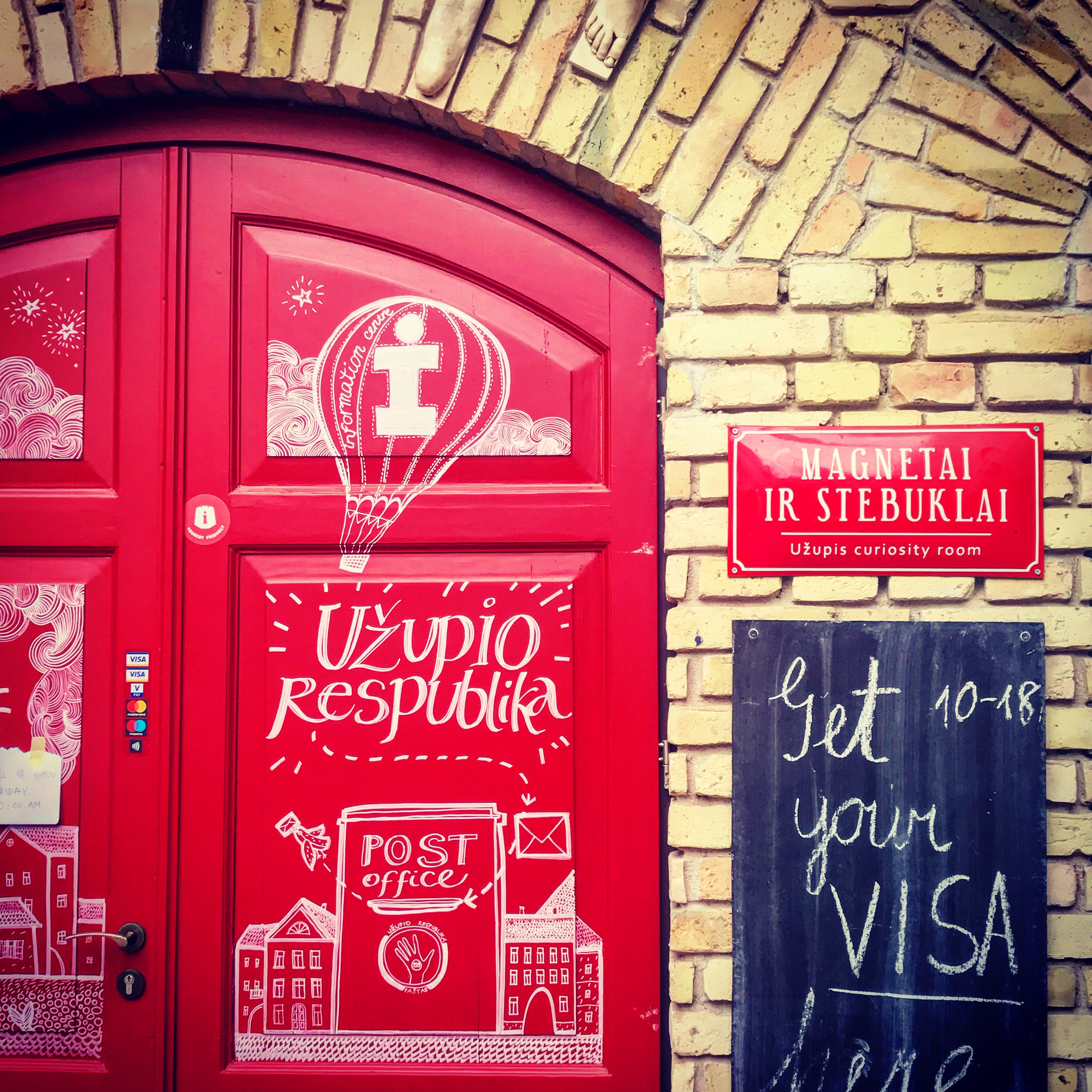 This image captures a vibrant red door featuring whimsical white illustrations and text, including "Uลพupio Respublika" and "Post Office," referencing the artistic micro-nation of Uลพupis. A sign on the brick wall reads "Magnetai ir Stebuklai" (Magnets and Wonders) and directs visitors to a curiosity room, while a chalkboard invites them to "Get your VISA here." The scene exudes a playful, creative atmosphere.