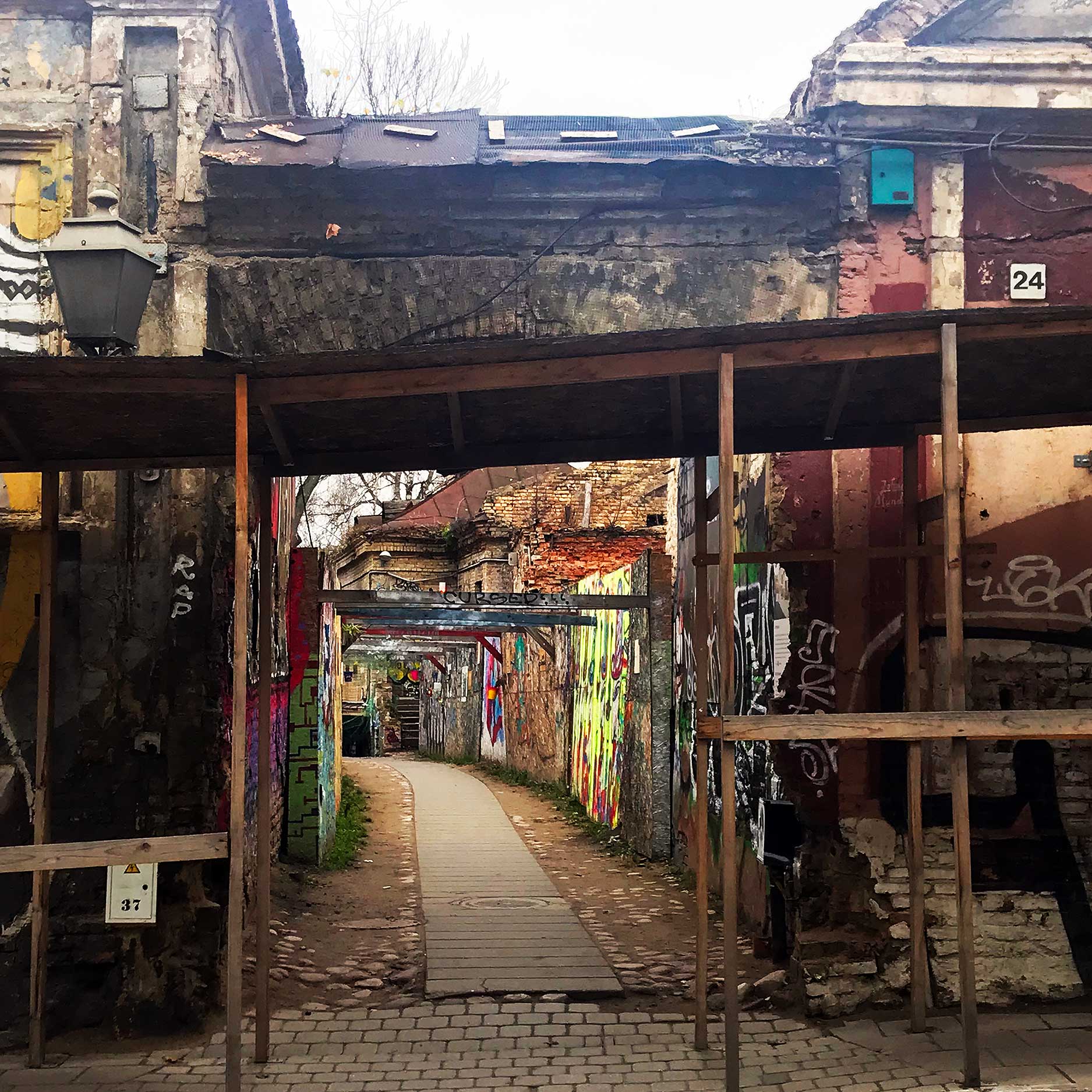 This image features a gritty urban alley framed by crumbling walls and scaffolding, with a path leading into a vibrant, graffiti-covered corridor. The juxtaposition of decayed architecture and colorful street art creates a raw and dynamic atmosphere. Numbered signs and rustic textures contribute to the charm of this artistic, yet industrial, space.