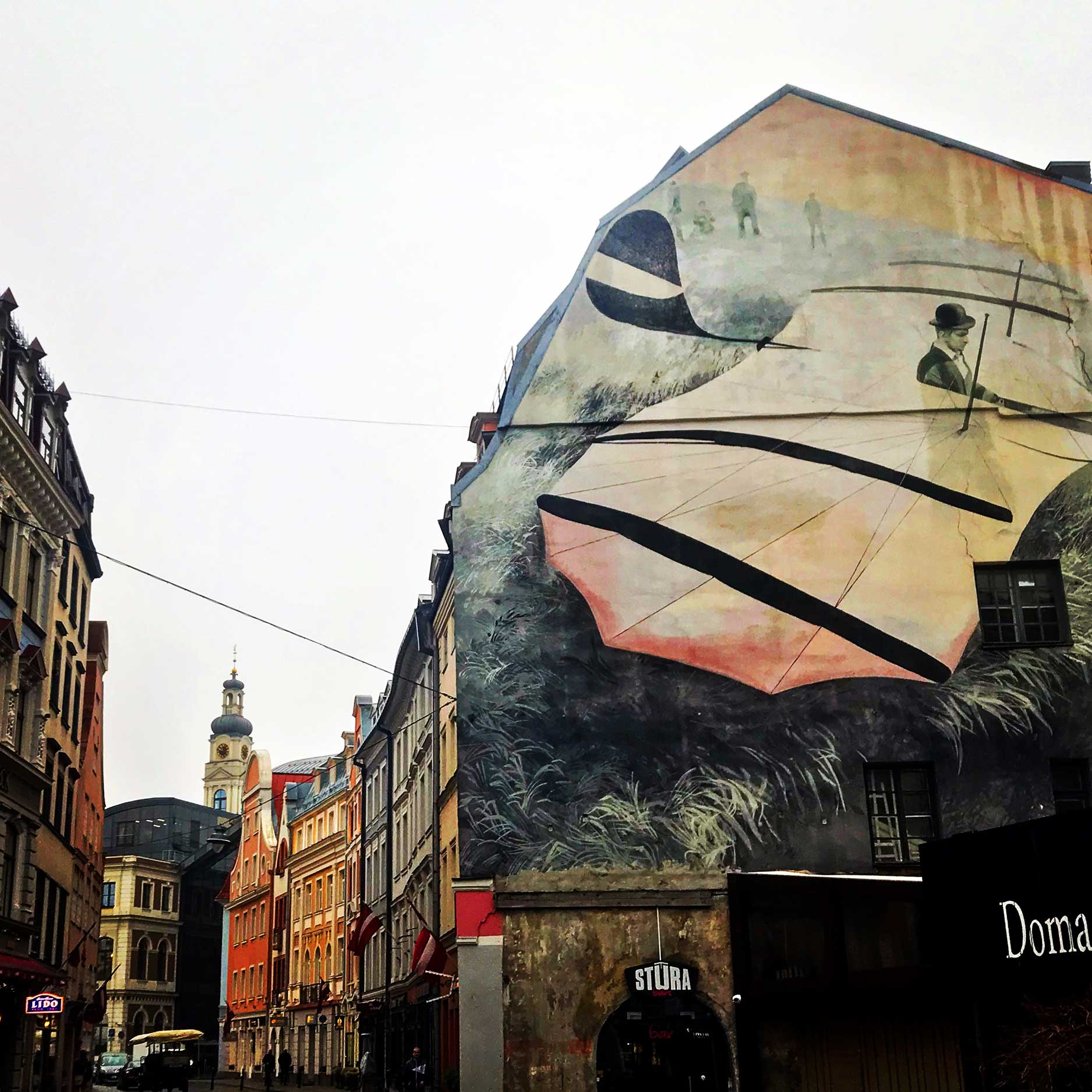 This image showcases a lively urban street lined with colorful historic buildings and a prominent mural painted on the side of one building. The mural depicts a surreal scene of a person with a bowler hat interacting with a large abstract structure resembling a kite or sail, set against a grassy background. In the distance, a tower with a golden spire adds a picturesque touch to the cityscape.