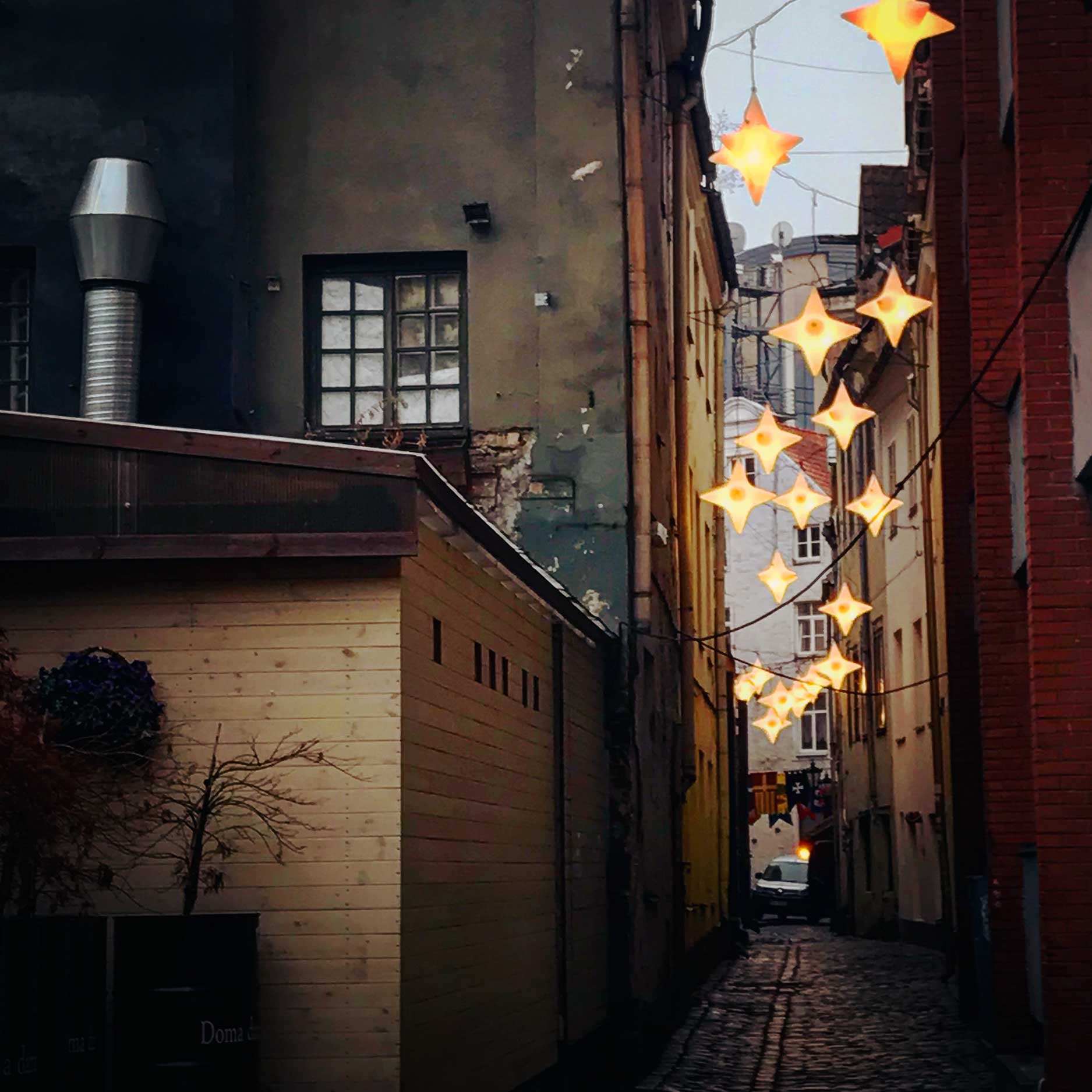 This image captures a narrow cobblestone alley adorned with glowing star-shaped string lights, creating a warm, festive atmosphere. The alley is framed by aged buildings with peeling paint and industrial details like a vent pipe, leading the eye toward a softly lit backdrop. The cozy yet urban charm makes it feel both intimate and inviting.