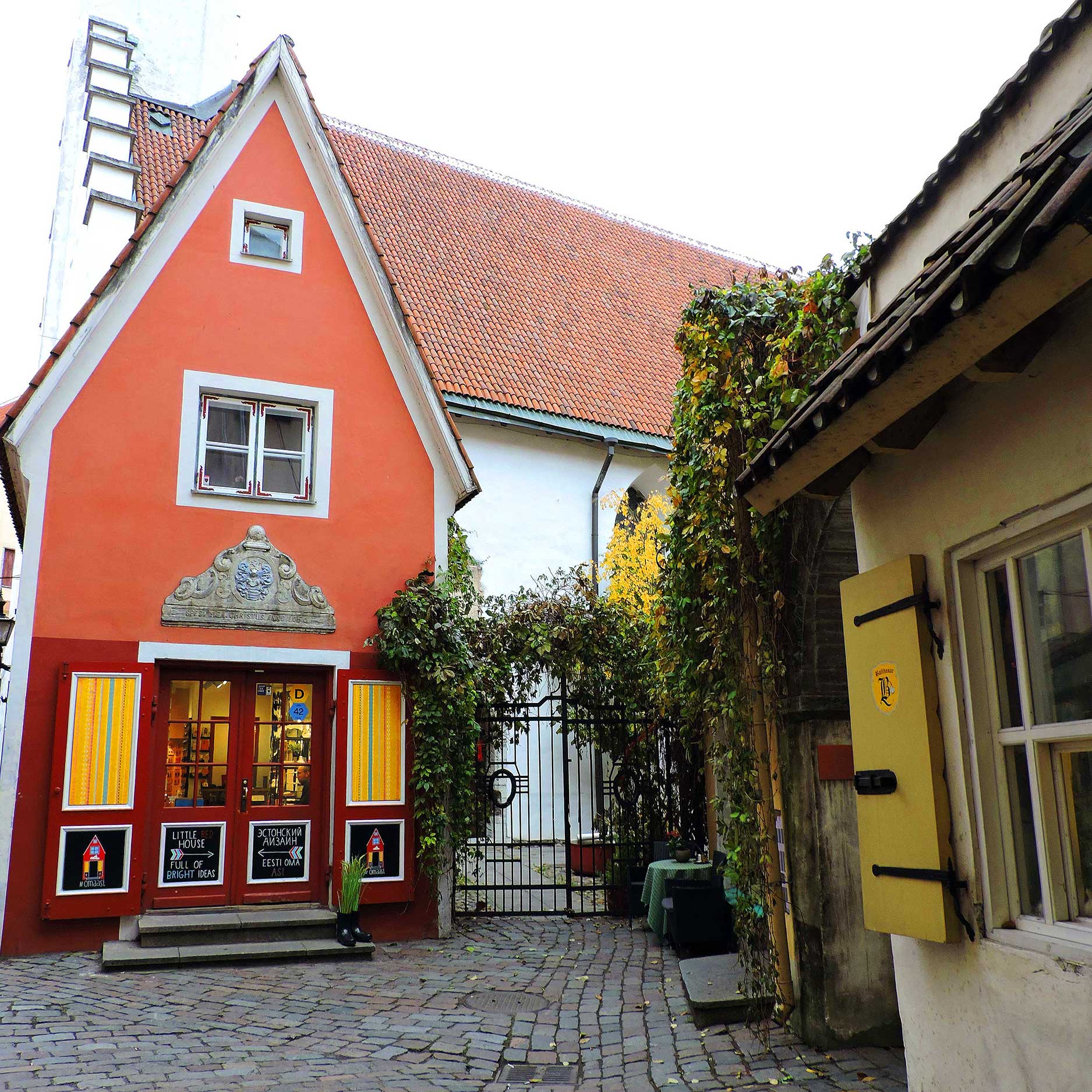 This image depicts a quaint cobblestone courtyard with a bright red house featuring yellow shutters and a decorative gable above the entrance. The windows showcase inviting displays, with signage reading โLittle House of Bright Ideas.โ Ivy-covered walls and a wrought iron gate add a touch of greenery and charm to this cozy, picturesque setting.