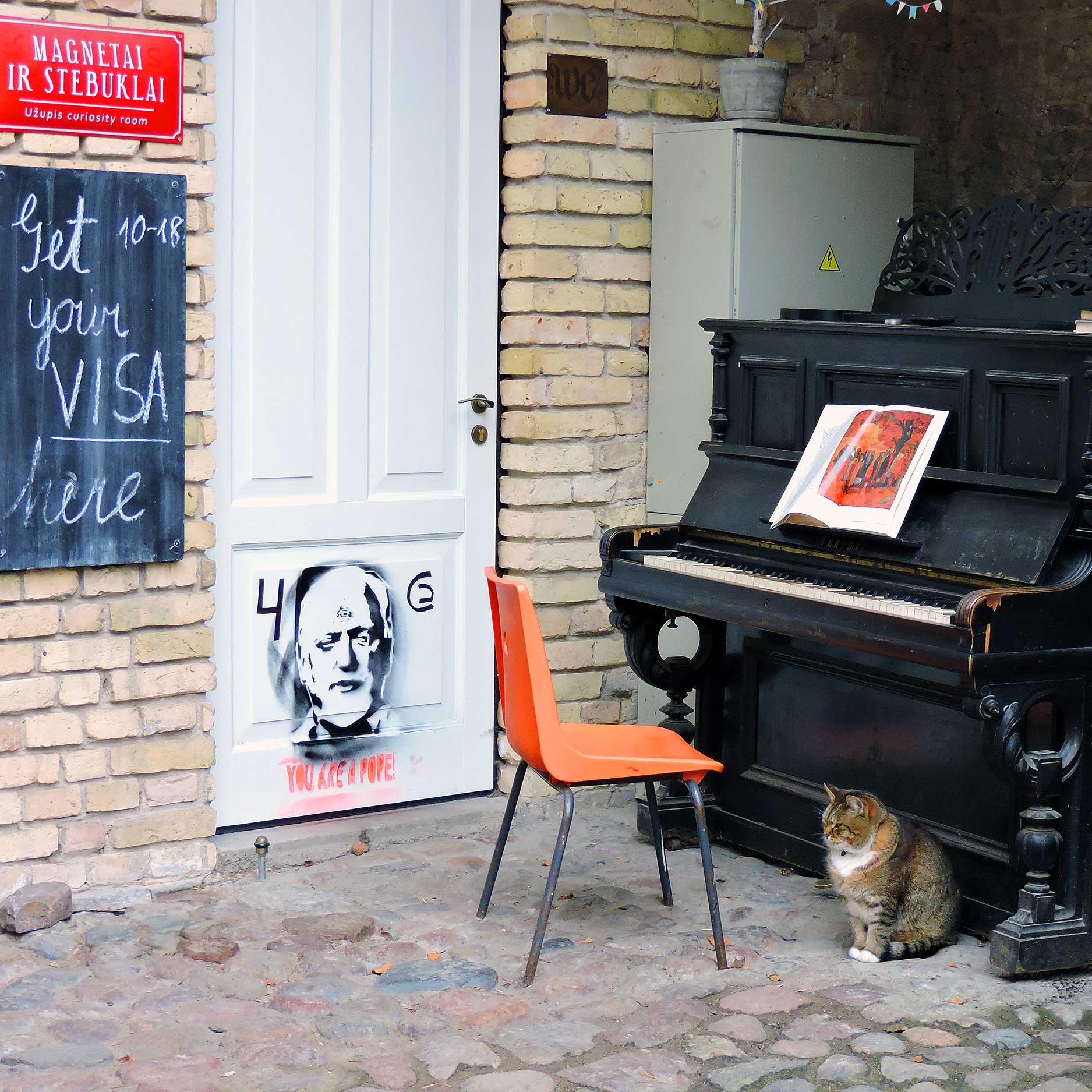 This image showcases a quirky outdoor scene featuring a white door with graffiti, including the phrase "You are a Pope," alongside a black upright piano with an open book displayed on top. An orange chair and a calm tabby cat sit on the cobblestone ground, creating a whimsical, artistic ambiance. A red sign reads "Magnetai ir Stebuklai," directing visitors to a curiosity room.