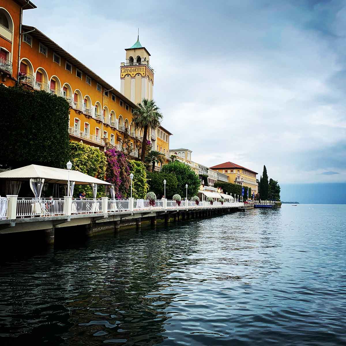 A picturesque view of the waterfront along Lake Garda, Italy, featuring the historic Grand Hotel with its distinctive yellow faรงade and clock tower. The hotel is adorned with lush greenery and vibrant bougainvillea climbing the walls, adding splashes of color. The promenade along the water is lined with elegant white railings and lamp posts, offering outdoor seating under canopies. The calm, reflective waters of Lake Garda extend into the distance under a cloudy sky, creating a serene and luxurious atmosphere.