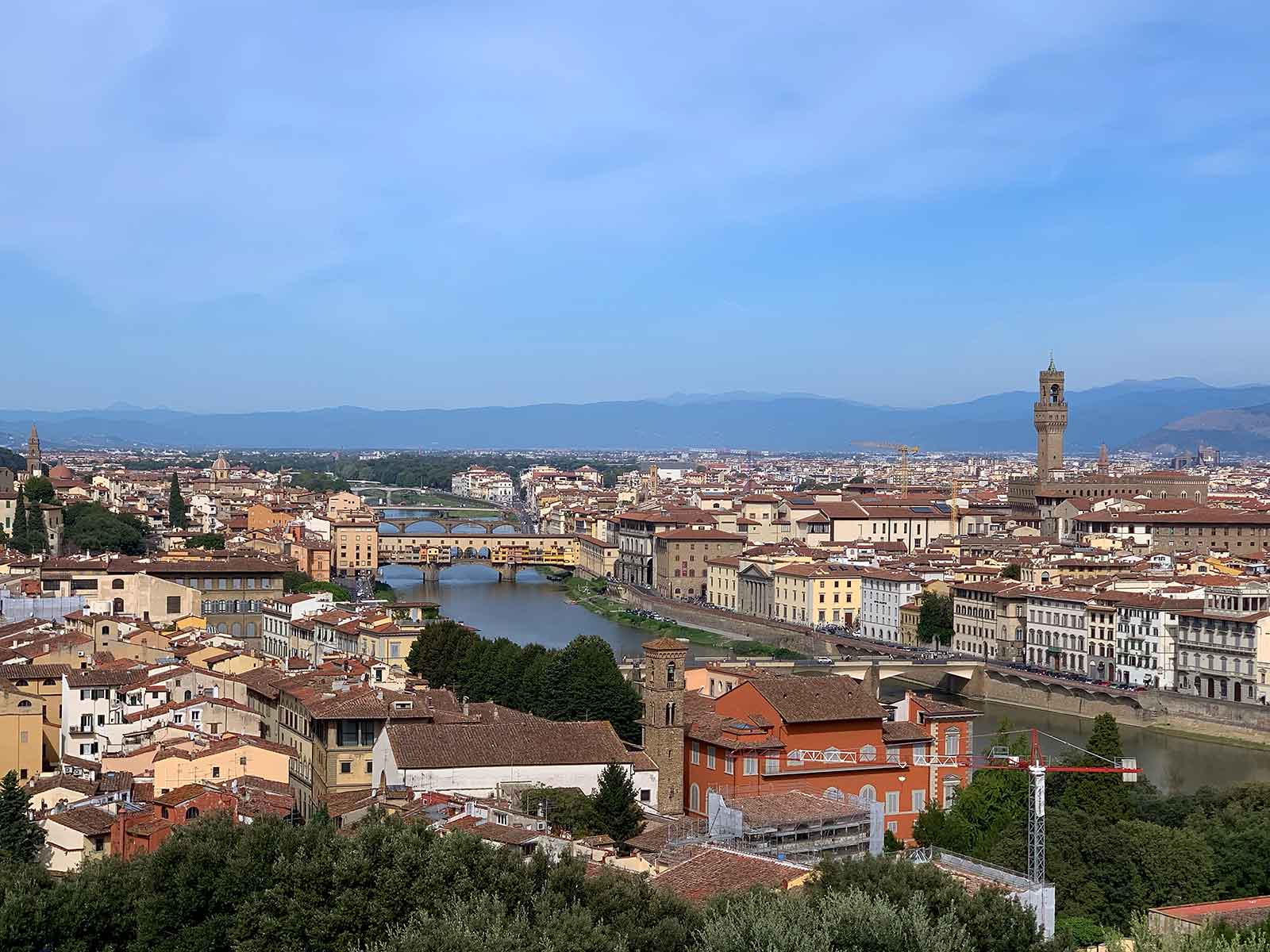 A panoramic view of Florence, Italy, showcasing the Arno River flowing through the city with several historic bridges, including the famous Ponte Vecchio, spanning its width. The city is dotted with red-tiled rooftops, historic buildings, and prominent landmarks, such as the Palazzo Vecchio with its tall tower rising above the skyline. In the background, the rolling hills and distant mountains frame the city, under a clear blue sky. The image captures the timeless beauty and architectural richness of Florence from an elevated vantage point.