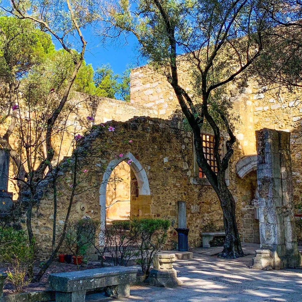 This image showcases the ruins within the grounds of Castelo de São Jorge in Lisbon, Portugal. Stone walls with arched doorways and scattered columns are shaded by tall trees, creating a serene and historic atmosphere under a bright blue sky. The play of light and shadows adds to the rustic charm of the ancient structure.