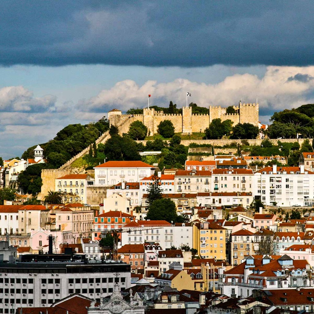 This image shows the iconic Castelo de São Jorge, a historic castle perched on a hilltop overlooking Lisbon, Portugal. The castle's stone walls and towers are surrounded by lush greenery, with the vibrant red-tiled roofs of the city's buildings in the foreground under a dramatic, cloudy sky.