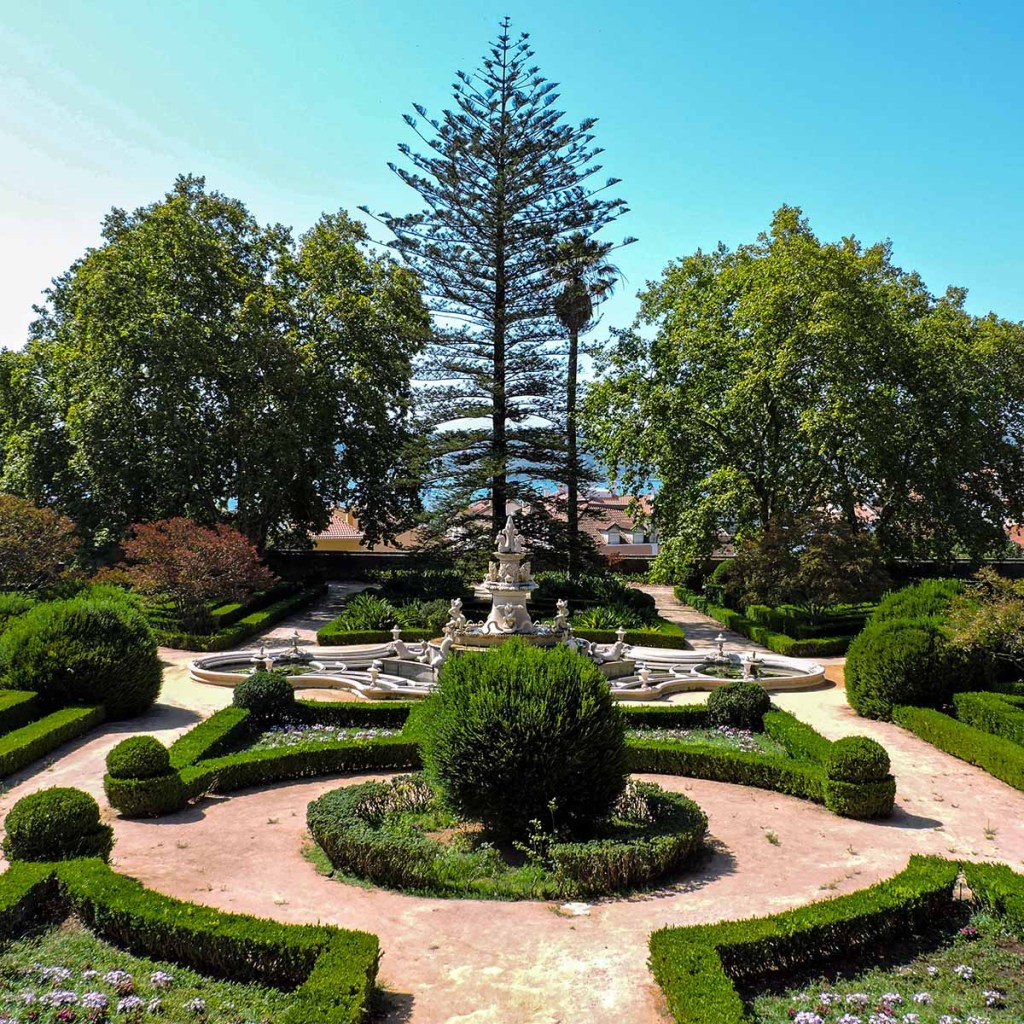 This image depicts a stunning formal garden in Lisbon, Portugal, featuring a central fountain surrounded by meticulously trimmed hedges and lush greenery. A towering tree dominates the backdrop, flanked by other mature trees that create a natural canopy over the garden. The symmetrical design, ornate fountain, and well-maintained paths evoke the elegance and tranquility of a classic European landscape.