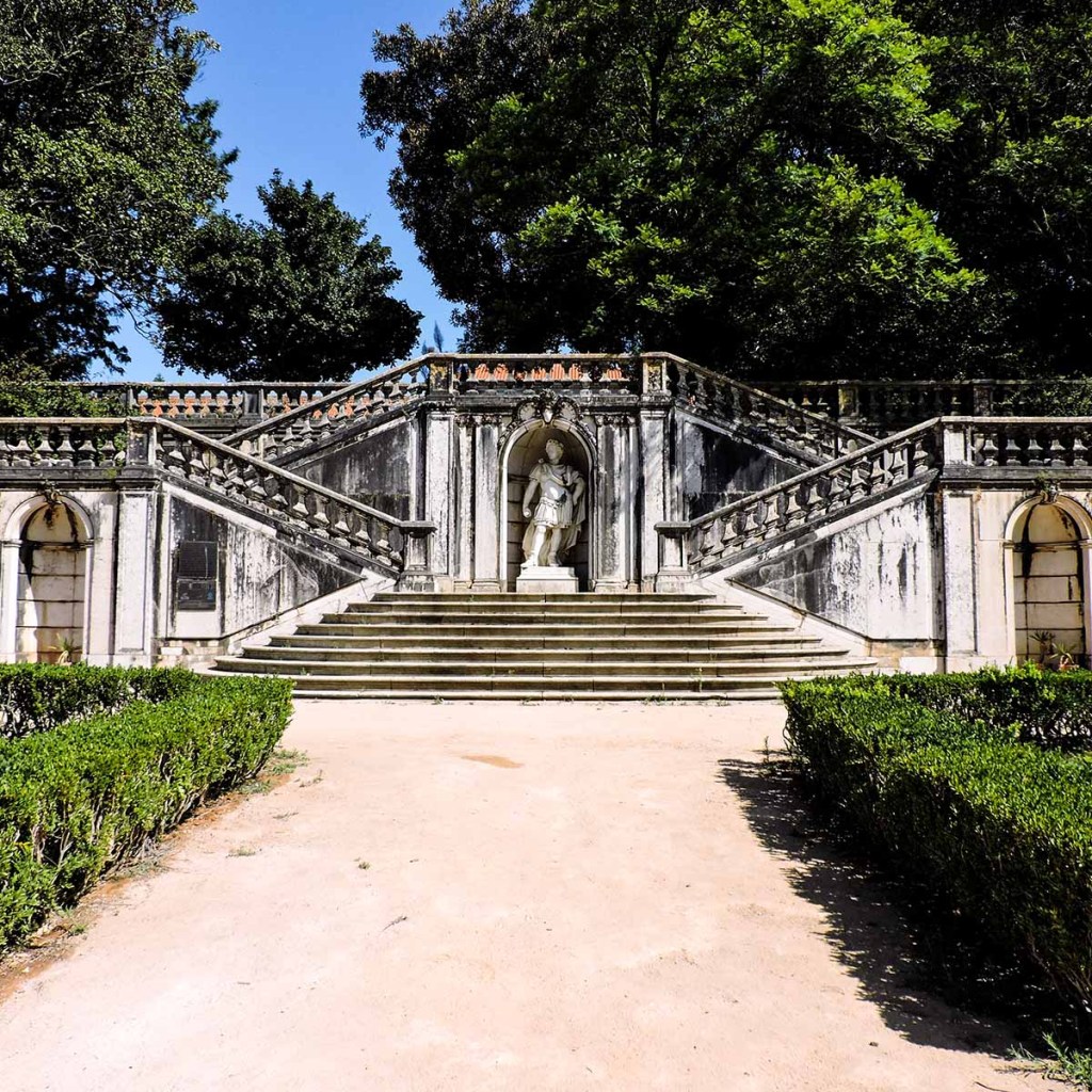 This image features a grand stone staircase in a historic garden in Lisbon, Portugal, leading up to a statue set within a niche. The staircase is framed by ornate balustrades and is surrounded by lush green hedges and towering trees, creating a stately and elegant atmosphere. The weathered appearance of the stone and the classical sculpture add to the timeless charm of this formal garden setting.