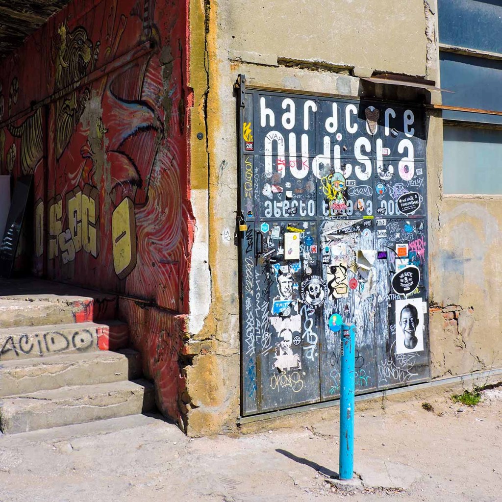 This image captures a gritty, urban scene in Lisbon, Portugal, featuring a wall covered in graffiti art and a heavily stickered door with the words “hardcore nudista” prominently displayed. The weathered building exterior, bold street art, and peeling posters create a raw, underground vibe. A blue bollard in the foreground contrasts with the rough textures, adding a splash of color to the otherwise industrial setting.
