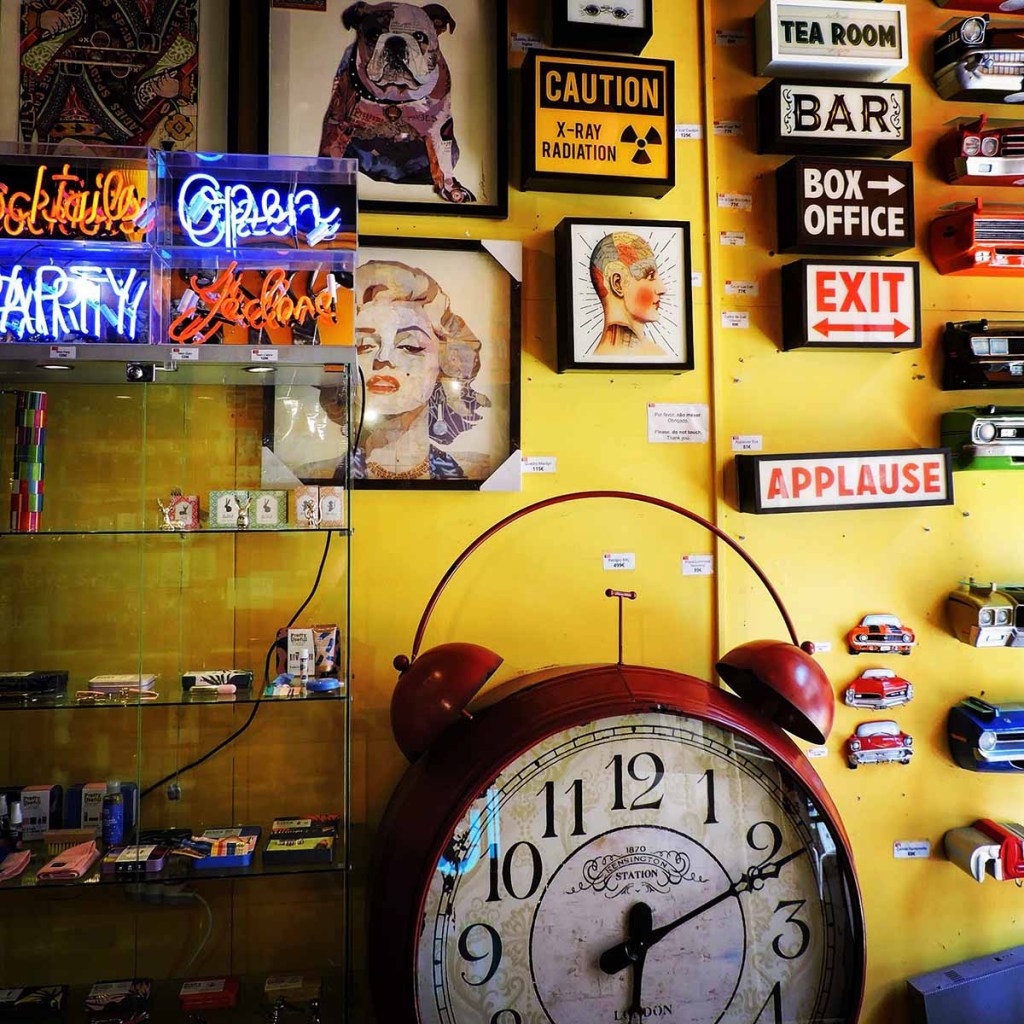 This image showcases an eclectic display wall inside a quirky shop or café, filled with vintage-inspired decor and signage. The vibrant yellow wall is adorned with neon signs, framed artwork, retro clocks, and a large decorative alarm clock. Playful elements like caution signs, a portrait of Marilyn Monroe, and various colorful retro memorabilia create a lively, nostalgic atmosphere.