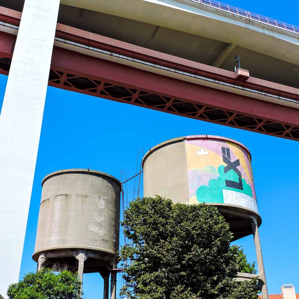 This image showcases two large, cylindrical water tanks beneath a bridge in Lisbon, Portugal, with one tank featuring colorful street art that reads "LX" against a backdrop of painted clouds. The tanks are positioned under the towering structure of the 25 de Abril Bridge, with its red metal supports and concrete pillars contrasting against the bright blue sky. The combination of industrial elements and urban art highlights Lisbon’s blend of modernity and creativity.