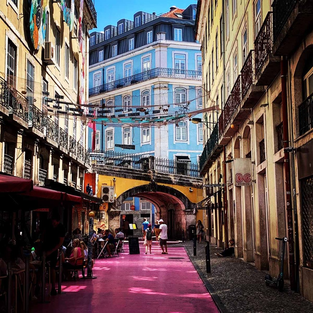 This image captures the vibrant Pink Street in Lisbon, Portugal, known for its lively atmosphere and colorful surroundings. The street is lined with historic buildings featuring wrought-iron balconies, with the striking pink pavement creating a playful contrast against the classic architecture. Overhead, strings of decorations and laundry add to the charming, bohemian vibe of this popular nightlife spot.