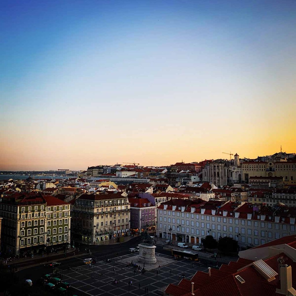 This image captures a panoramic view of Lisbon, Portugal, at sunset, with the city’s red-roofed buildings bathed in the soft, warm glow of the fading light. The scene includes a spacious square with a prominent statue in the center, framed by historic architecture and distant hills. The sky transitions from a deep blue to a golden hue, adding a serene and picturesque ambiance to the cityscape.