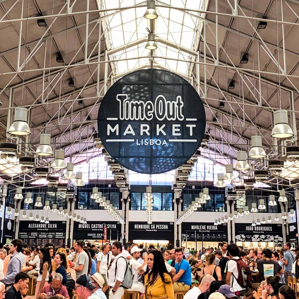 This image captures the bustling interior of the Time Out Market in Lisbon, Portugal, filled with people enjoying the vibrant atmosphere. The large, iconic black and white Time Out Market sign hangs prominently from the ceiling of the spacious, industrial-style market hall, surrounded by food stalls from renowned chefs. The lively crowd and stylish lighting fixtures highlight the market's dynamic blend of gourmet food and social space.