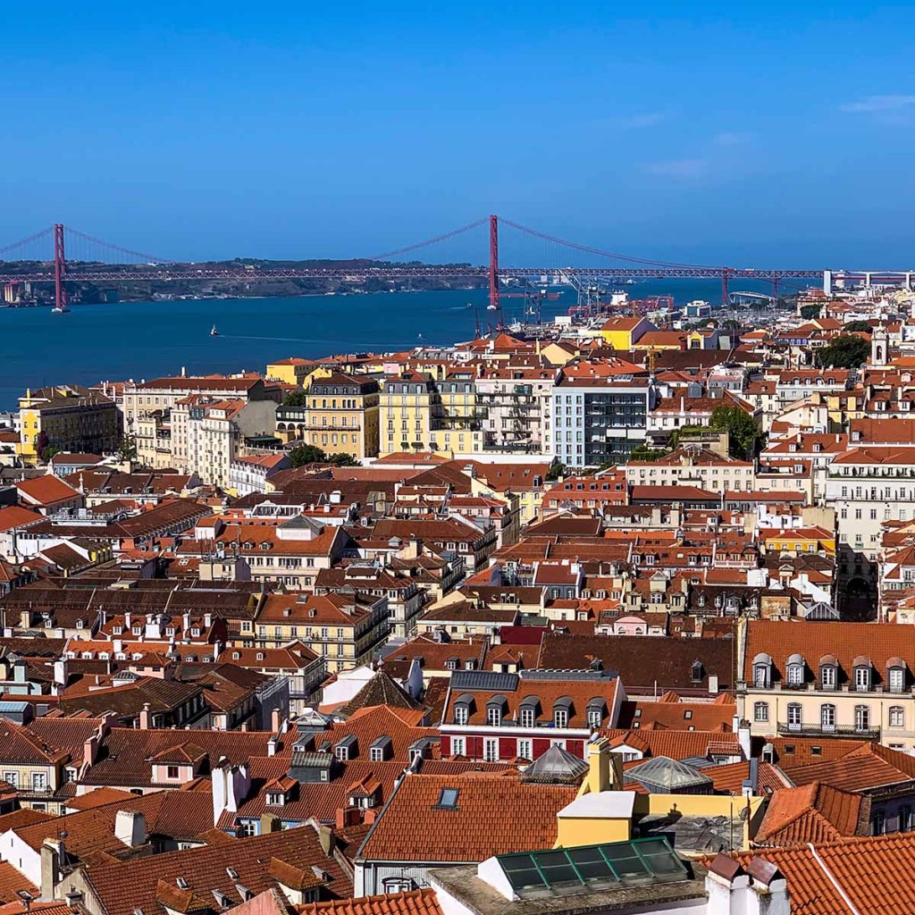 This image captures a scenic view of Lisbon, Portugal, with its iconic red-tiled rooftops densely packed across the city. In the background, the red 25 de Abril Bridge spans the Tagus River under a clear blue sky, adding to the city’s picturesque coastal charm.