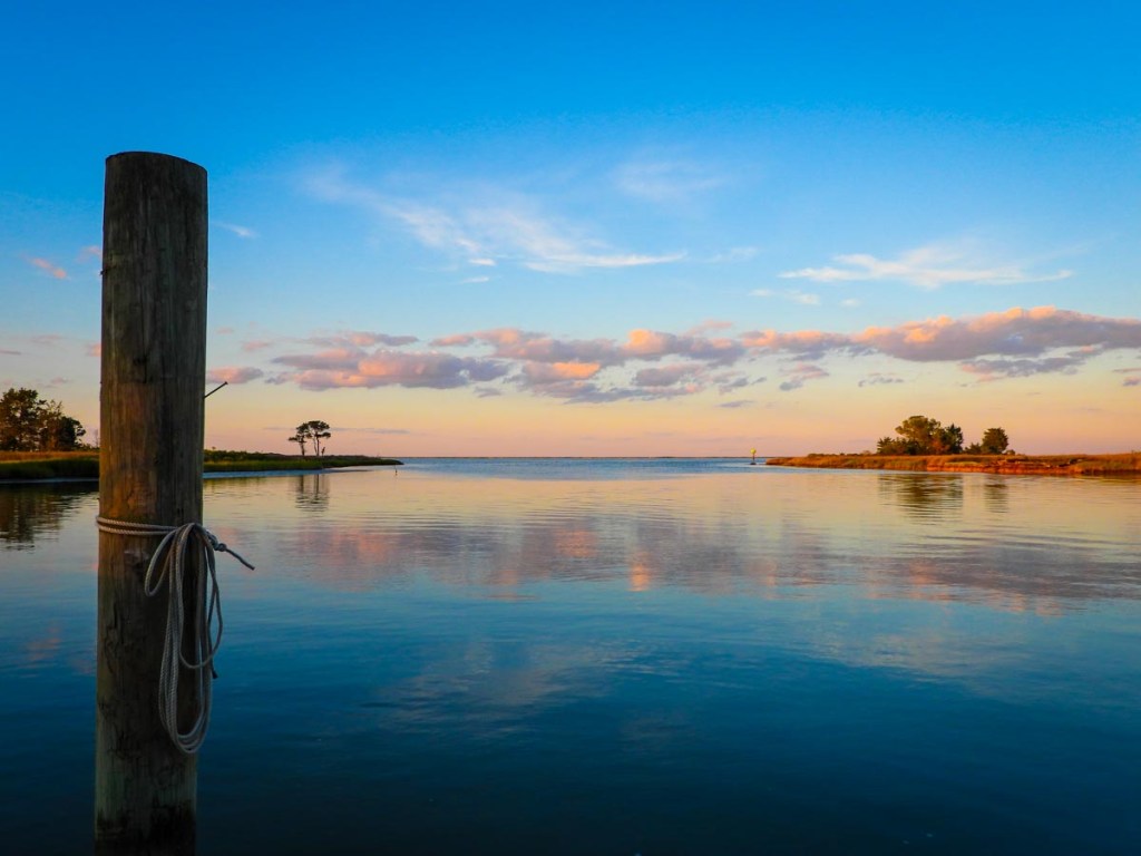 A peaceful waterside scene at sunset, featuring a wooden post with a coiled rope in the foreground and a serene expanse of water reflecting the colorful sky. In the distance, a small tree-lined island and a vivid horizon complete the picturesque view.