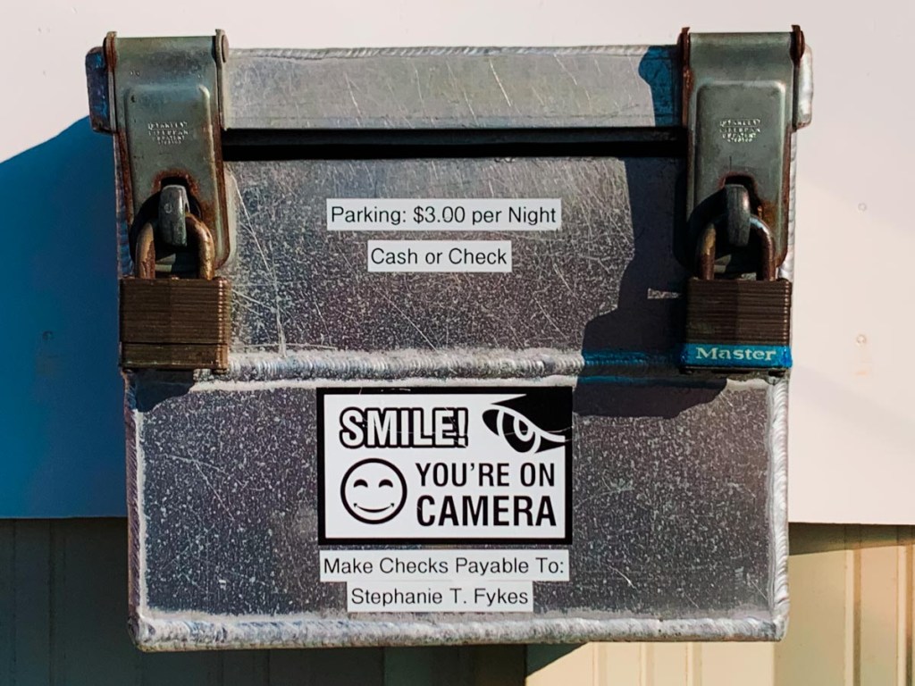 A metal payment box with two padlocks has several signs attached. One sign reads "Parking: $3.00 per Night, Cash or Check," another says "SMILE! YOU'RE ON CAMERA" with a smiley face and an eye graphic, and a third instructs "Make Checks Payable To: Stephanie T. Fykes." The box is used for collecting parking fees.