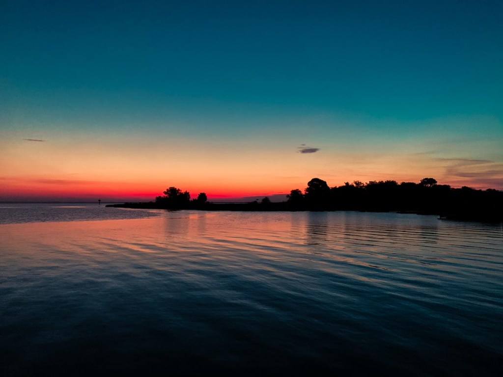 A stunning sunset over a calm body of water, with the sky transitioning from deep blue to vibrant shades of orange and pink near the horizon. Silhouettes of trees and landforms are visible in the distance, adding to the tranquil and picturesque nature of the scene.