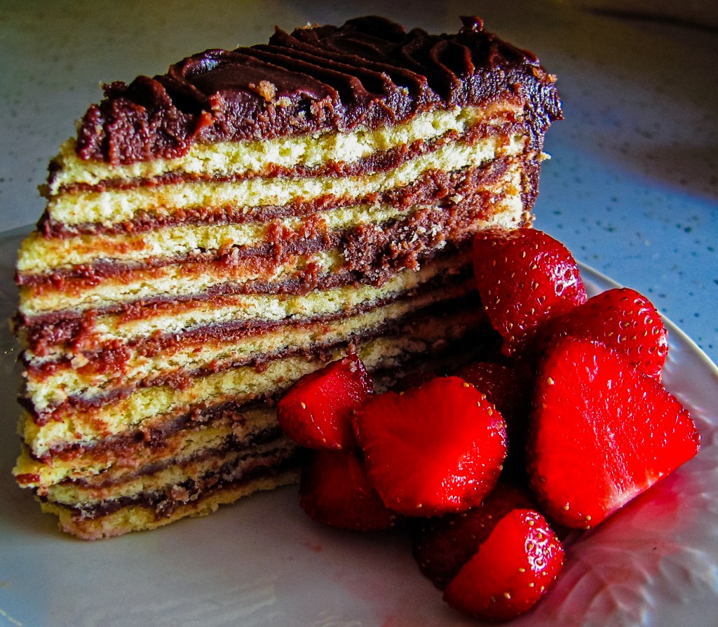 A slice of layered chocolate smith island cake with rich frosting sits on a plate, accompanied by fresh, juicy strawberry halves. The cake features multiple thin layers of sponge cake and chocolate filling, creating a visually appealing and appetizing dessert.