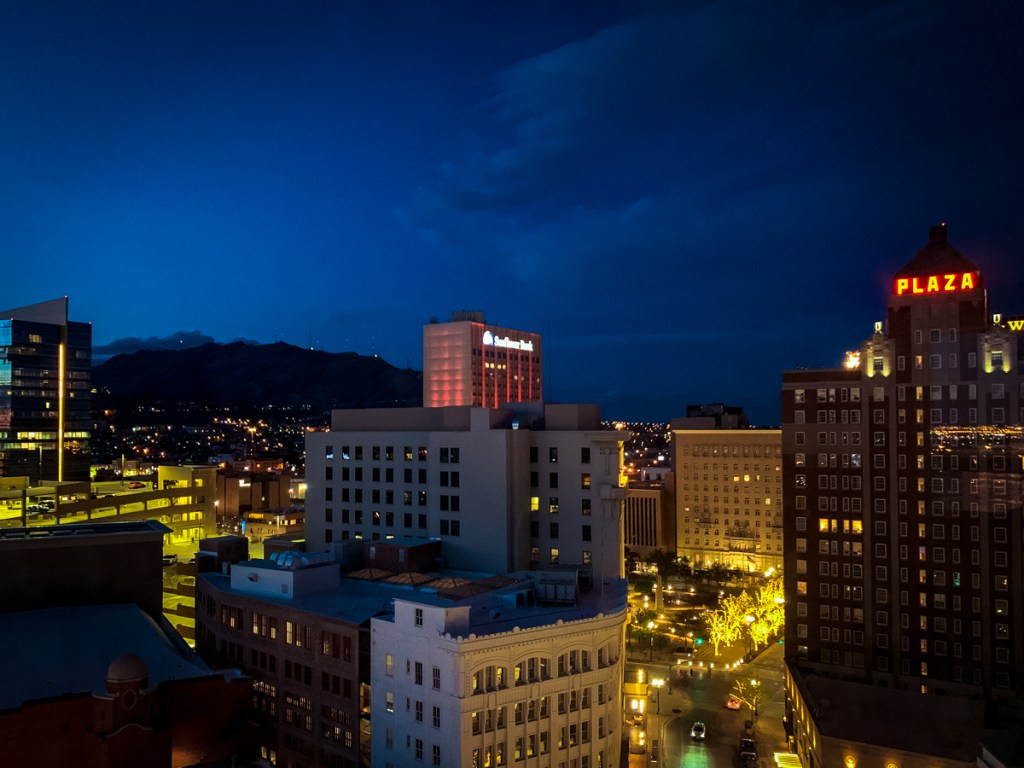 A nighttime cityscape showcases brightly lit buildings in downtown El Paso, with the "Plaza" sign glowing in red on top of one of the tallest buildings. The streets below are illuminated with warm yellow lights, and the horizon reveals a dark mountain range against the deep blue sky following sunset. The scene captures a lively, yet serene atmosphere, with a mix of modern and historic architecture visible. Distant city lights twinkle across the landscape, adding depth to the urban view.