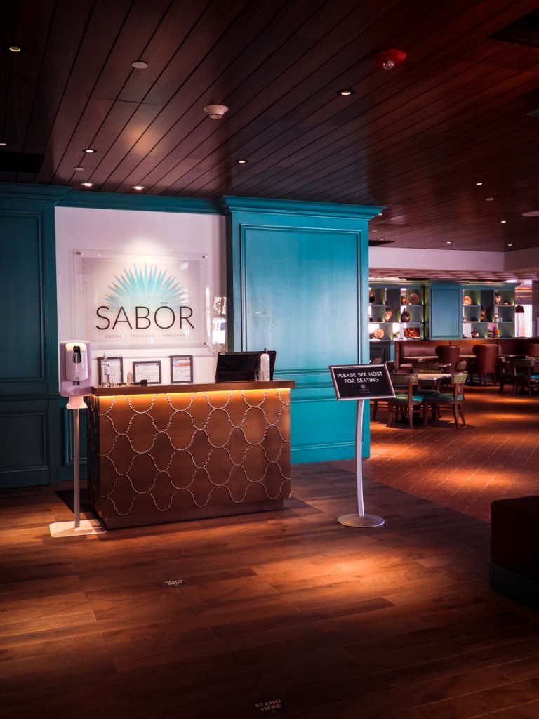 This image shows the entrance area of a restaurant named "Sabor," with its logo displayed on the wall behind a reception desk. The desk features a sleek design with a brown pattern, and a sign next to it reads, "Please see host for seating." The lighting is warm, with a modern, cozy atmosphere visible in the background, including teal-colored walls and decor.