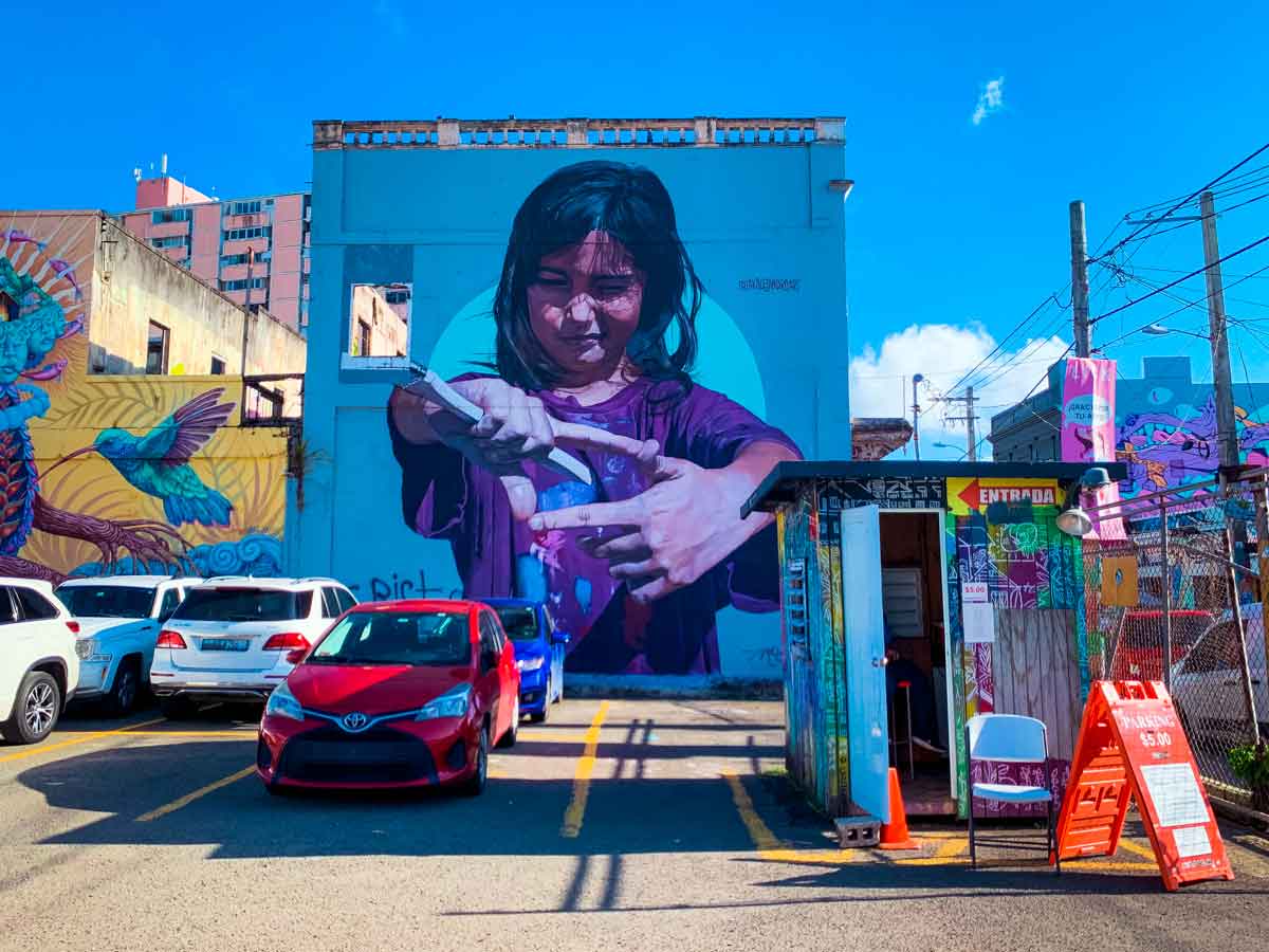 This image depicts a colorful urban scene dominated by a large mural of a young girl on the side of a building. The mural shows the girl intensely focused as she carefully pours liquid from one container into another, her expression and the detailed painting capturing a moment of concentration. The mural is set against a bright blue background, with the word "ENTRADA" (meaning "entrance" in Spanish) visible on a sign nearby, indicating the entrance to a parking area. The foreground includes several parked cars, with other vibrant murals visible on adjacent buildings, including a hummingbird and an abstract creature, contributing to the lively and artistic atmosphere of the area. The bright, clear sky adds to the vibrant, energetic feel of the scene.