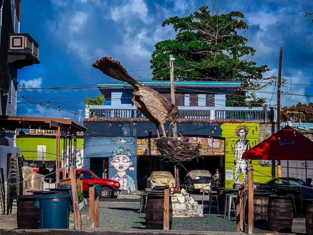 This image captures an outdoor scene featuring a large sculptural installation of a bird perched in a nest, made from what appears to be wood or metal. The sculpture is centrally located in a small open space surrounded by buildings with colorful murals, including one of a person with a halo-like effect around their head on the right side. A bright blue sky with scattered clouds provides a vibrant backdrop, and there are strings of lights hanging across the area, adding a festive atmosphere.