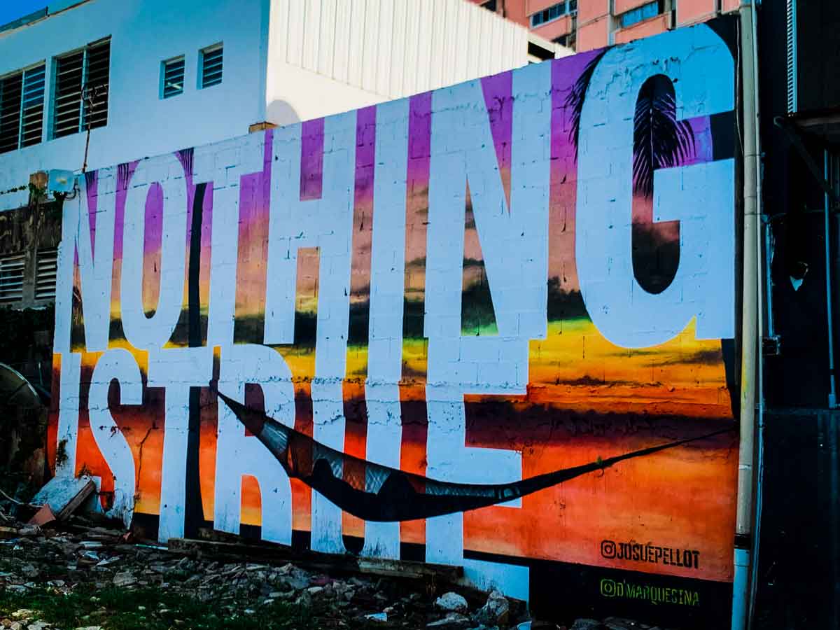 This image features a mural with the large, bold text "NOTHING IS TRUE" painted over a vibrant, sunset-colored background. The background includes hues of orange, yellow, purple, and black, resembling a tropical sunset with palm trees silhouetted against the sky. The text takes up most of the wall, with the words "NOTHING" and "TRUE" in stark white, making a strong visual impact. Below the mural, the Instagram handles "@josuepellot" and "@marquesina" are visible, likely crediting the artists. A hammock is also strung across the mural, adding an element of contrast between the permanence of the art and the temporary, relaxed nature of the hammock. The scene is set in an urban area with some debris in the foreground, suggesting a blend of art and everyday life.