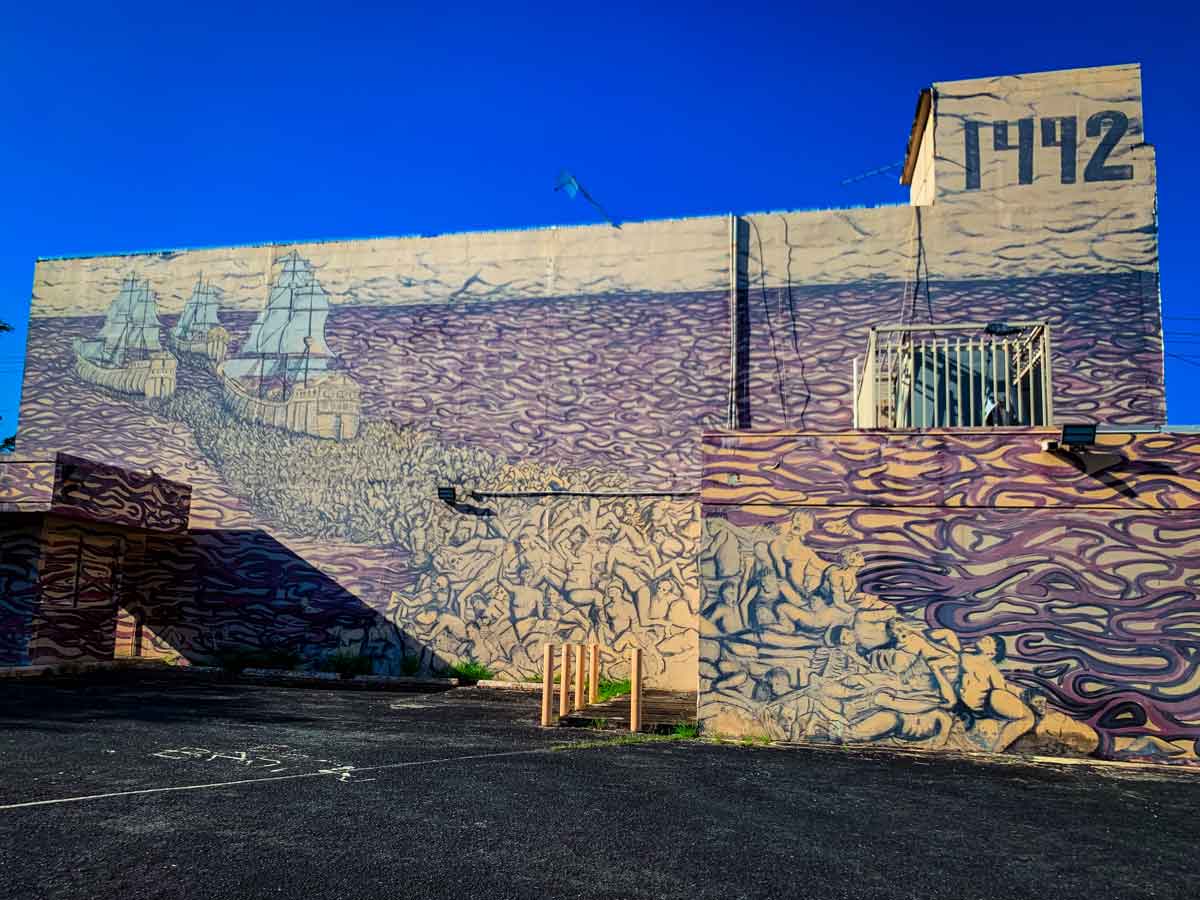 This image shows a large mural on the side of a building, depicting a historical scene of the journey to puerto rico, with three sailing ships approaching land, accompanied by a mass of human figures that seem to flow like waves onto the shore. The scene appears to reference the year "1492," which is prominently displayed in the upper right corner, likely alluding to the arrival of Christopher Columbus in the Americas. The artwork is rendered in shades of beige and purple, with intricate details in the figures and waves, creating a dramatic and powerful visual narrative. The mural covers the entire side of the building, with some portions wrapping around corners, emphasizing the expansive and immersive nature of the scene. The deep blue sky contrasts sharply with the muted tones of the mural, making the artwork stand out even more.