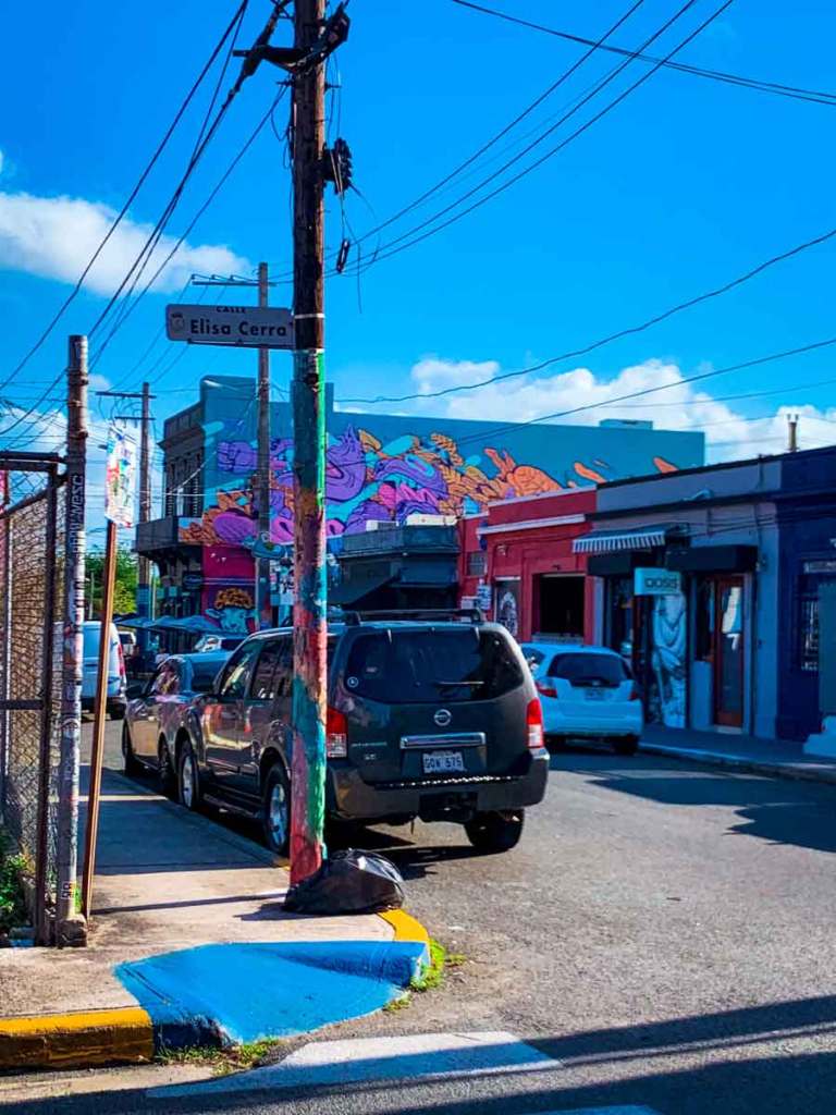 This image shows a lively street scene with colorful buildings and murals. The street is identified by a sign as "Calle Elisa Cerra." In the distance, a large mural on the side of a building features abstract, brightly colored shapes in pink, purple, orange, and blue, adding a vibrant touch to the neighborhood. Several cars are parked along the street, and utility poles are adorned with stickers and paint, contributing to the urban and artistic atmosphere. The clear blue sky and scattered clouds provide a cheerful backdrop, enhancing the colorful and dynamic feel of the area.