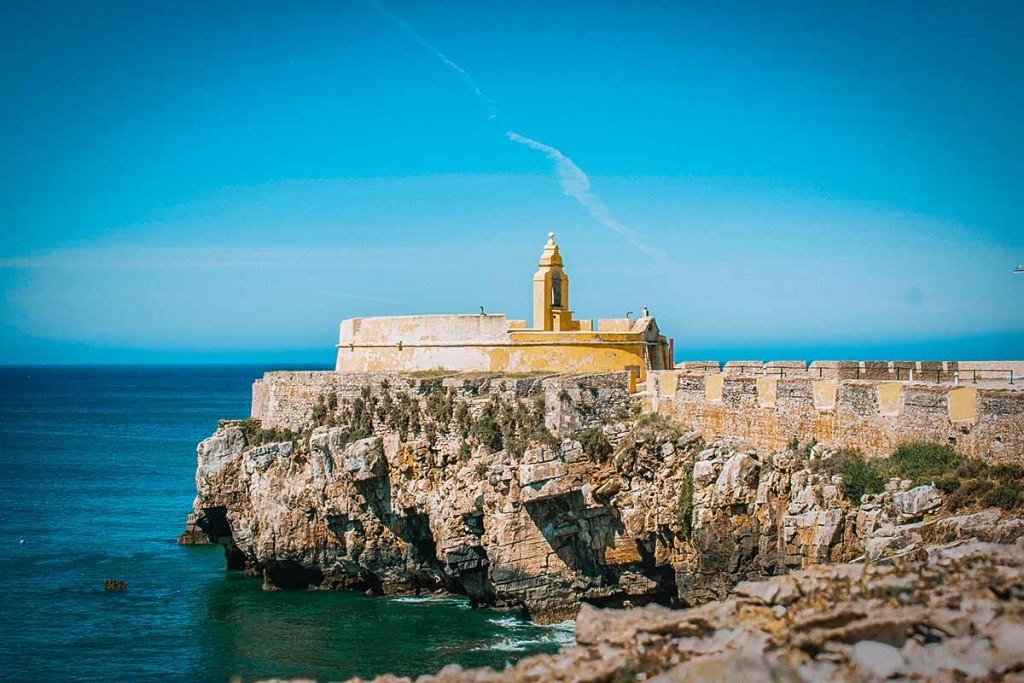 This image showcases a bright yellow fortress perched on rocky cliffs overlooking the ocean. The vibrant blue sky and sea create a striking contrast with the historic structure, making it a perfect coastal highlight.