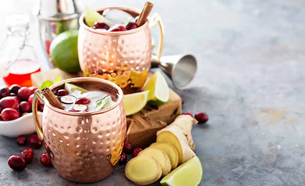 Two copper mugs filled with festive Friendsgiving drinks —cranberry Moscow mules garnished with cranberries, lime wedges, mint leaves, and cinnamon sticks. Surrounded by fresh ginger slices, lime, and cranberries, these drinks bring a cozy and refreshing flavor to any Friendsgiving gathering.