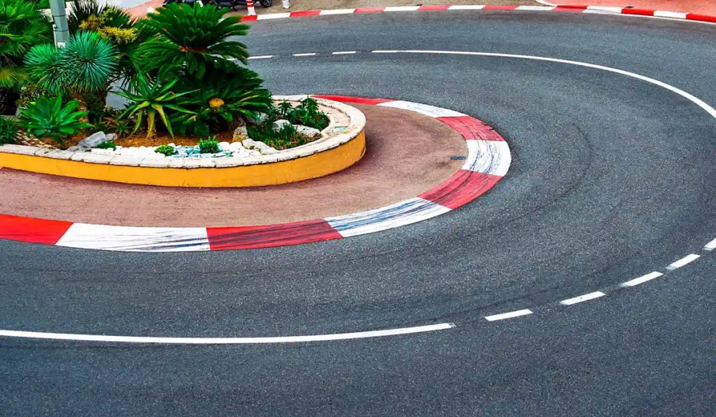 This image depicts a section of a race track with a sharp curve, bordered by red and white curbs. In the center of the turn, there is a landscaped island featuring various green plants and small rocks. The track surface appears smooth, indicating a well-maintained racing circuit.