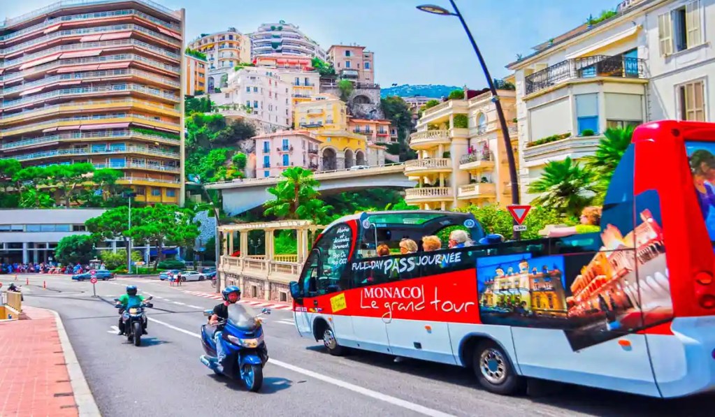 This image shows a bustling street scene in Monaco with a sightseeing bus labeled "Monaco Le Grand Tour" in the foreground. The bus is filled with tourists, and nearby, two motorcyclists are riding on the road. In the background, a hillside is dotted with colorful buildings and lush greenery, highlighting Monaco's vibrant urban landscape.