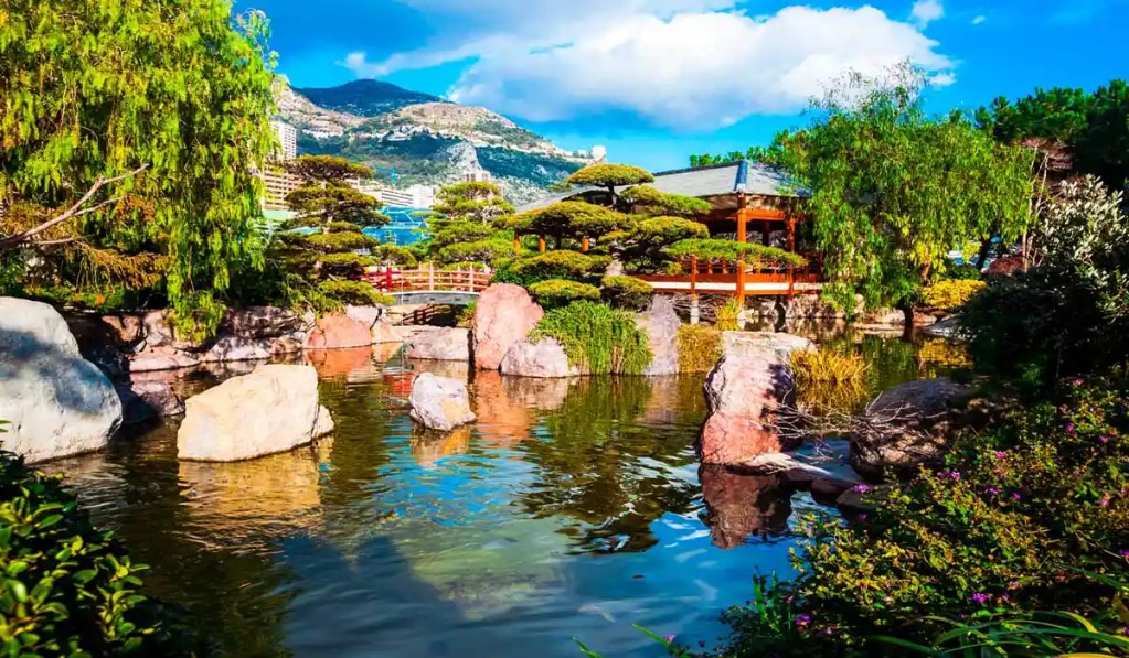 This image captures the serene beauty of the Japanese Garden in Monaco. The scene features a tranquil pond surrounded by well-manicured trees, large rocks, and lush greenery. A traditional Japanese-style pavilion and bridge are visible, adding to the garden's authentic and peaceful ambiance. The backdrop includes mountainous terrain and blue skies, enhancing the picturesque setting.