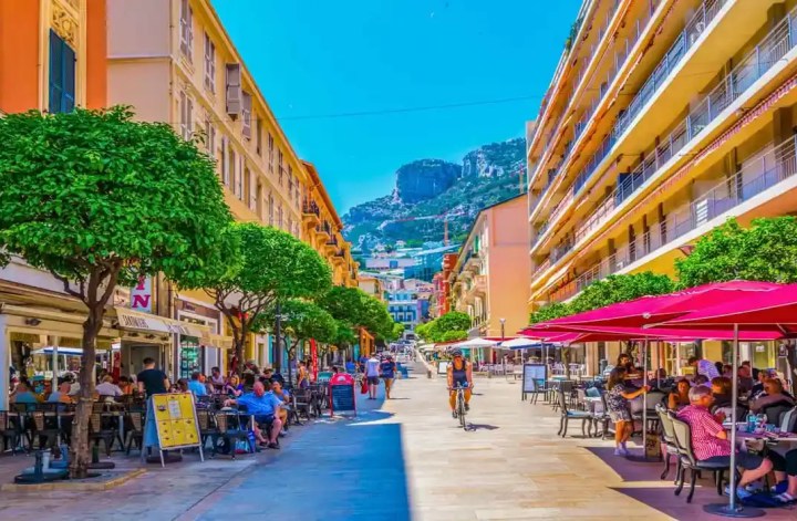 This image shows a vibrant street in Monaco lined with outdoor cafes and restaurants, where people are dining and socializing. The street is flanked by tall buildings and lush, well-manicured trees, with a backdrop of mountains under a clear blue sky. Bright red umbrellas provide shade for the patrons, adding a splash of color to the lively scene.