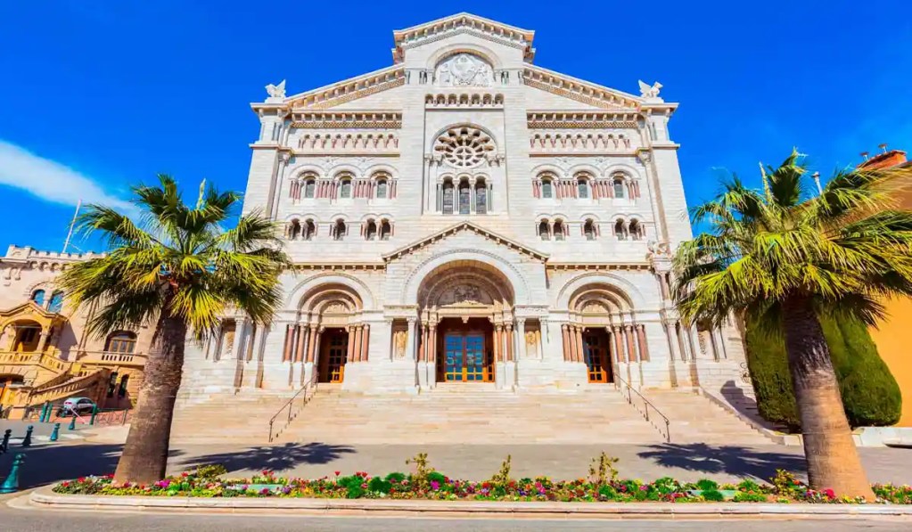 On the top of the list of things to do in Monaco. This image depicts the Saint Nicholas Cathedral in Monaco, a stunning example of Romanesque-Byzantine architecture. The cathedral's grand facade features intricate stone carvings, arched windows, and a rose window above the main entrance. Flanked by palm trees and a colorful flower bed, the building stands out against the clear blue sky, creating a striking and picturesque scene.