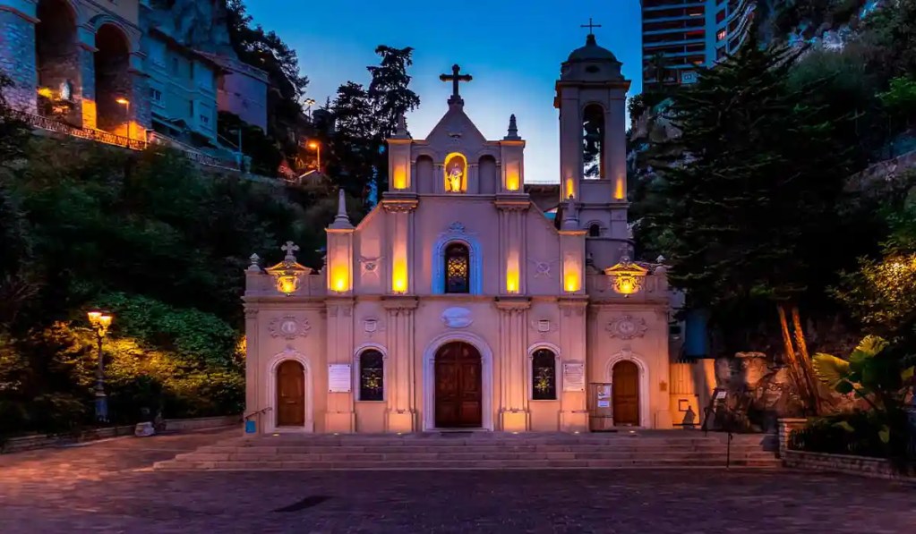 This image shows the Sainte-Dévote Chapel in Monaco illuminated at dusk. The small, historic chapel features a symmetrical façade with arched windows and doors, highlighted by soft lighting that accentuates its architectural details. The chapel is surrounded by lush greenery and set against a backdrop of hillside buildings, creating a serene and picturesque scene.
