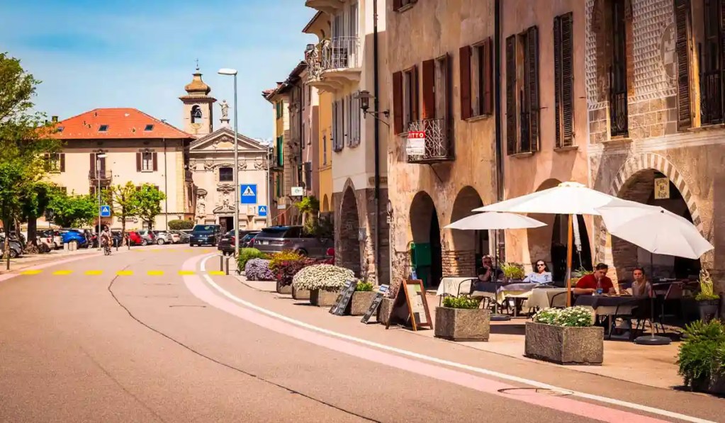 A charming street scene in Bissone, Switzerland, featuring outdoor cafรฉ seating with white umbrellas along a quaint cobblestone road lined with historic buildings and colorful flowers. A church with a distinctive bell tower is visible in the background, adding to the village's picturesque atmosphere.