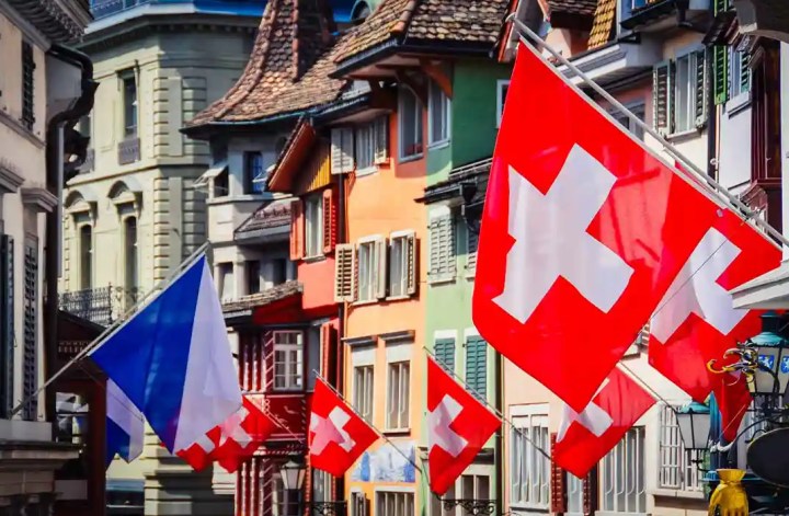 This image shows a street in Zurich, Switzerland, adorned with several Swiss flags featuring white crosses on red backgrounds. The buildings lining the street are painted in various vibrant colors, adding to the festive atmosphere. The scene captures the charm and lively spirit of the city's old town.