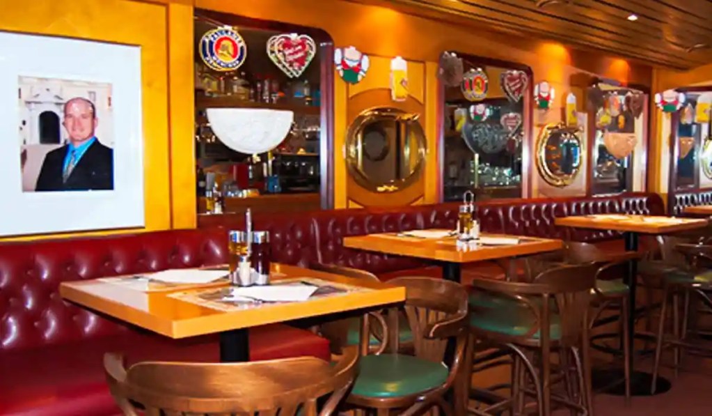 The cozy interior of a restaurant with wooden tables and chairs, red leather banquette seating, and warm yellow walls. Decorative mirrors, framed photos, and traditional heart-shaped decorations adorn the walls, adding to the inviting and intimate atmosphere.