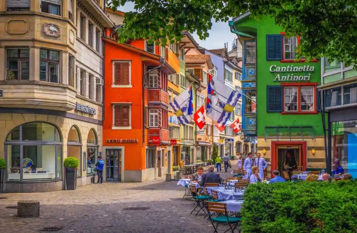 This vibrant image depicts a picturesque street in Zurich, Switzerland, lined with colorful buildings and outdoor café tables. The scene includes a green building with the sign "Cantinetta Antinori" and a bright orange building housing "Vero Moda." Flags hang across the street, adding to the lively and charming atmosphere.