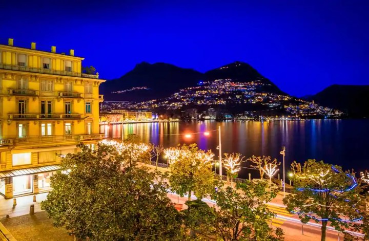 A vibrant nighttime scene in Lugano, Switzerland, showcasing a beautifully illuminated lakeside with twinkling lights reflecting on the water. The foreground features a grand building and tree-lined street adorned with festive lights, while the background reveals the silhouette of mountains dotted with sparkling lights from the hillside homes.